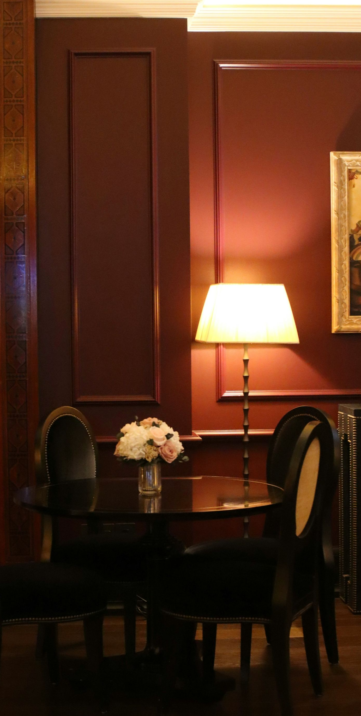 A small table with a floral centerpiece surrounded by four chairs in a dimly lit room with dark red walls and a lamp.