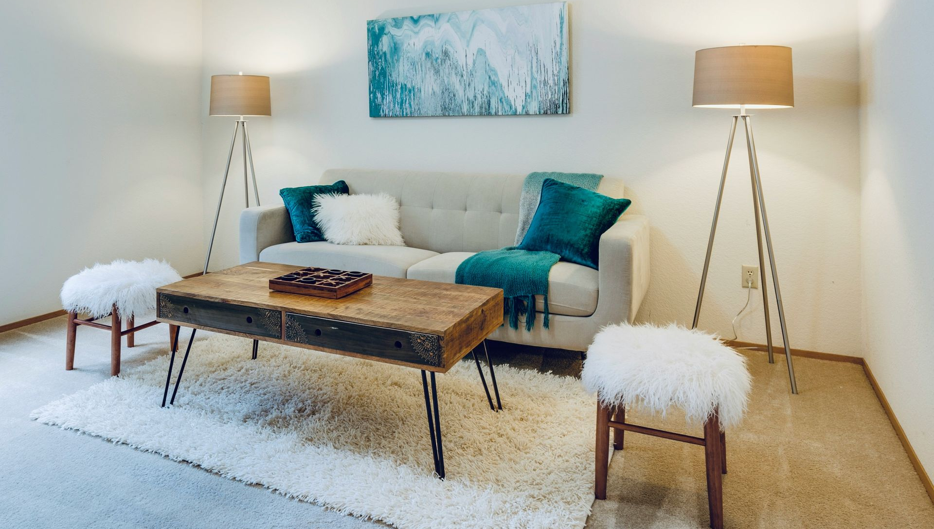 Living room with beige sofa, turquoise accents, two tripod lamps, a rustic coffee table, and fluffy stools.