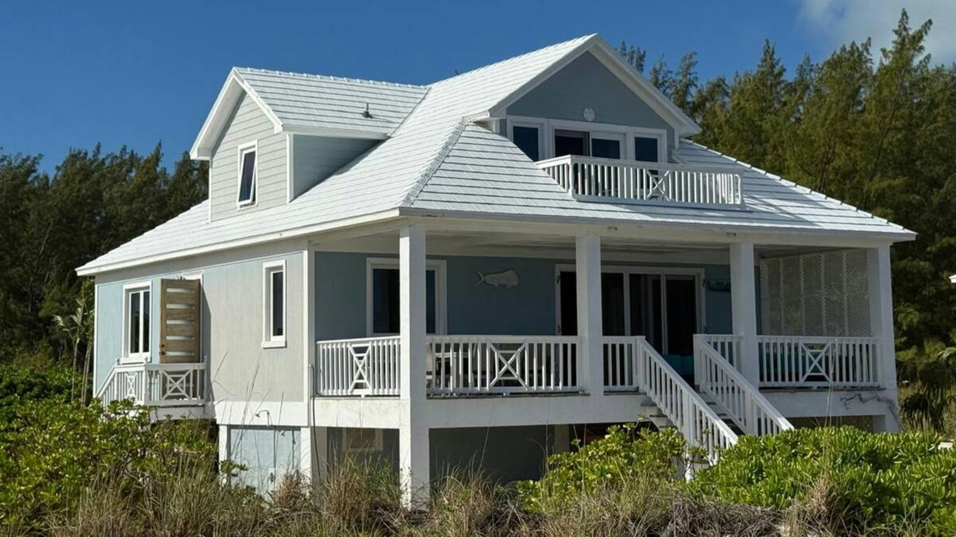 A light blue, two-story beach house on stilts with a white roof and wrap-around porch, surrounded by green foliage.