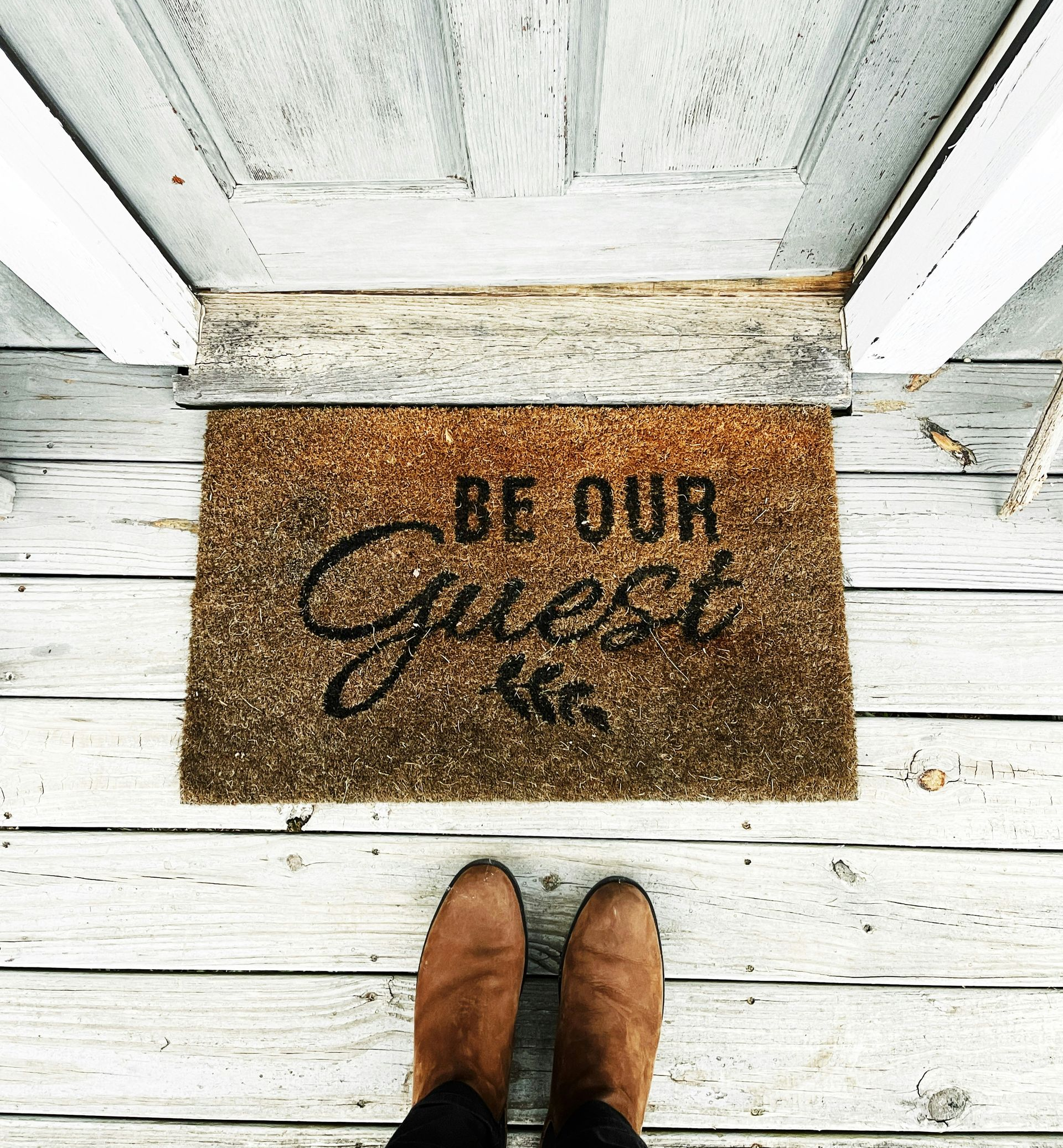 A person wearing brown boots stands on a porch in front of a welcome mat that says, 