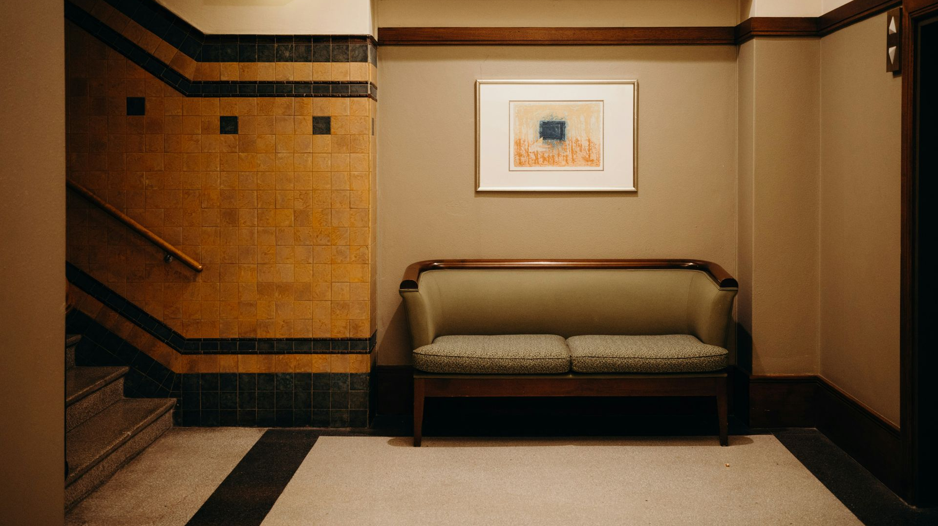 A hallway with tan walls, a framed abstract print, a green settee, and stairs against a brown-tiled wall with an exit sign.