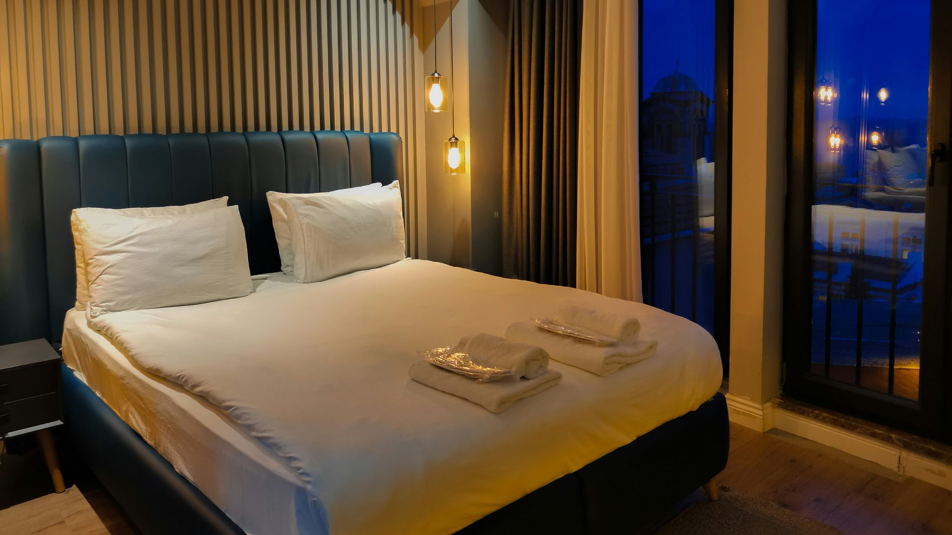 A modern bedroom with a blue tufted headboard, white bedding, and tall windows looking out at dusk.