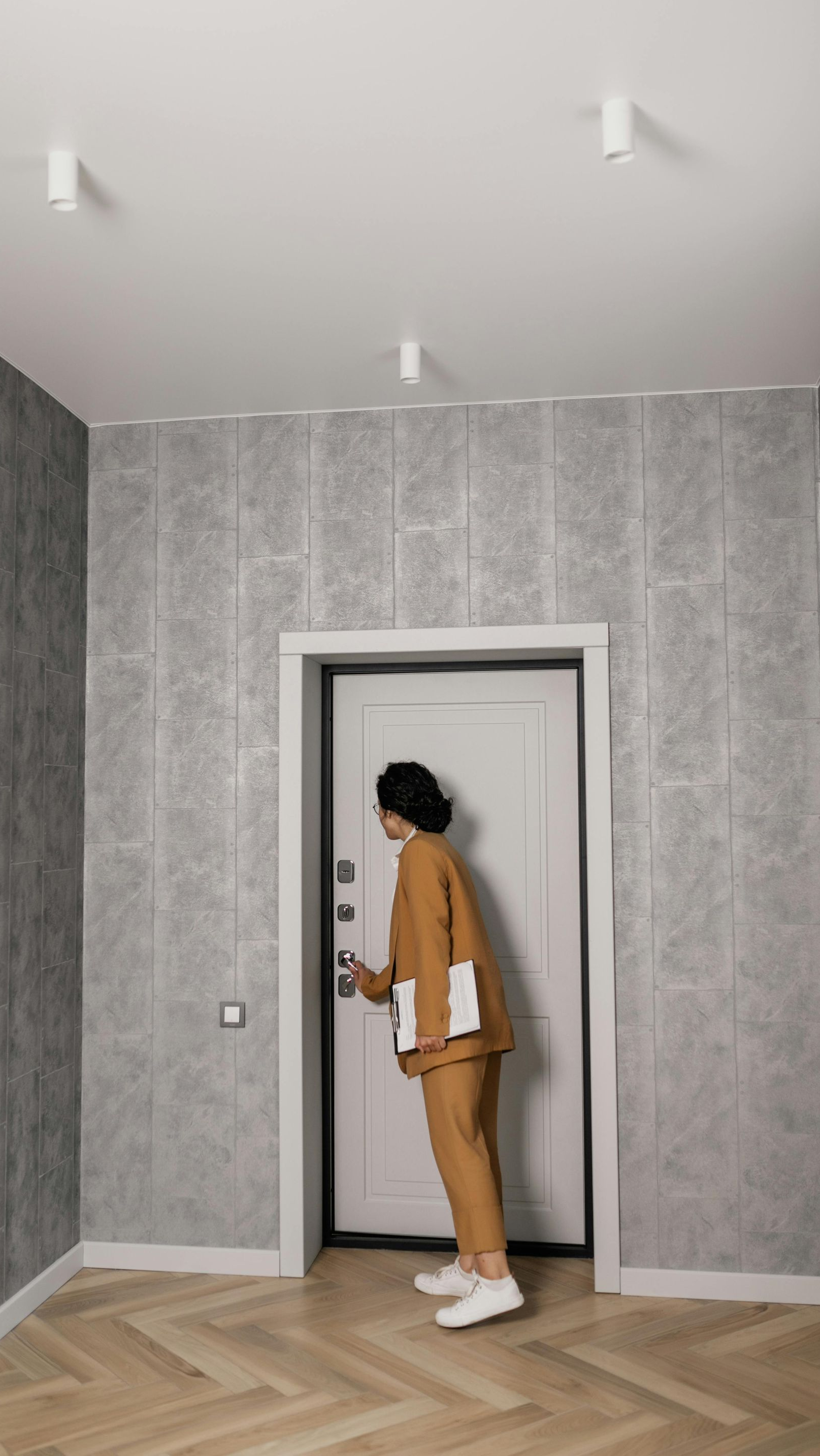Person in a tan suit opening a white door, holding documents, in a room with grey wallpaper and wood floor.