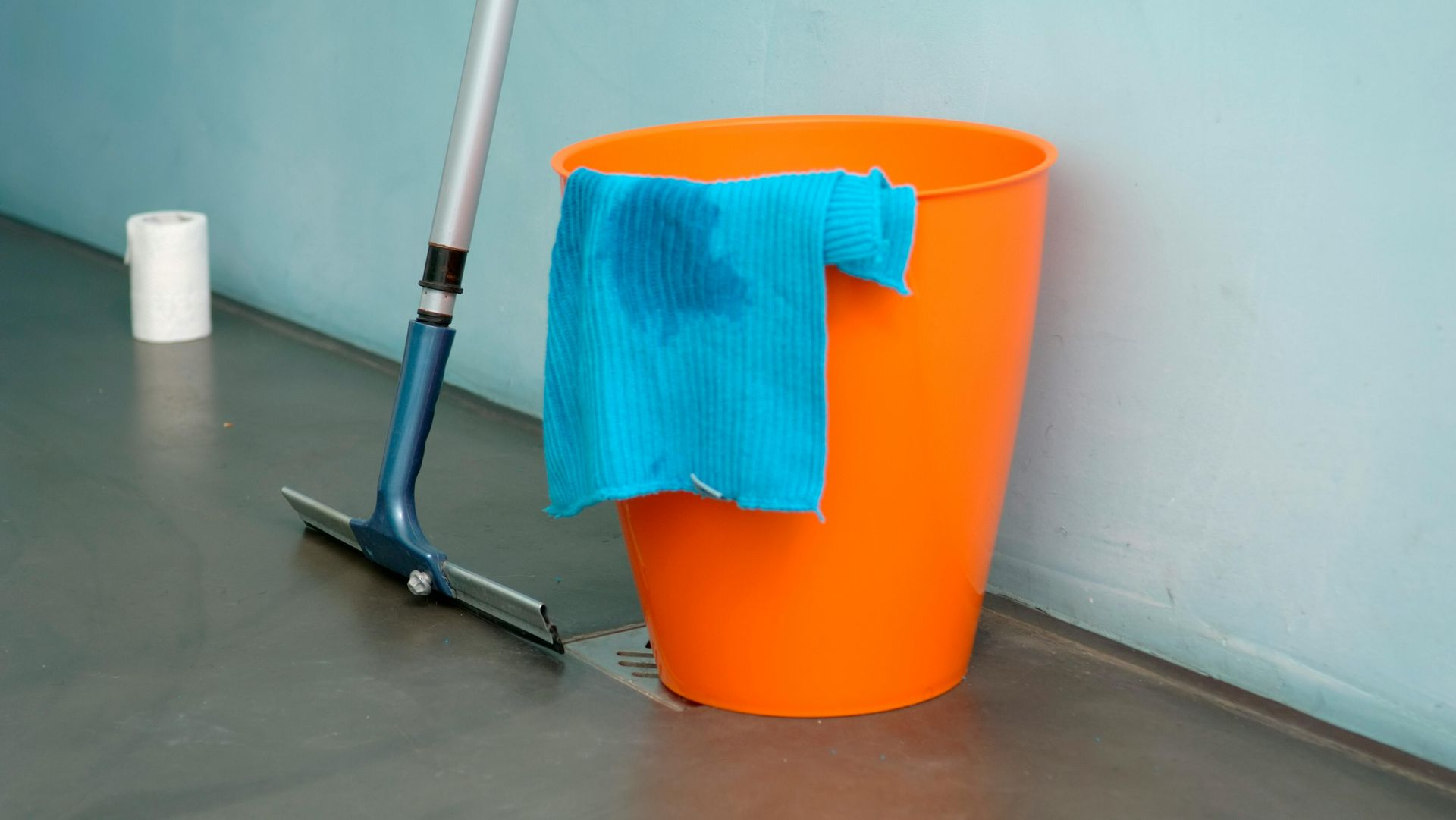 An orange bucket with a blue cloth draped over it sits on a grey floor next to a squeegee and a roll of paper towels.