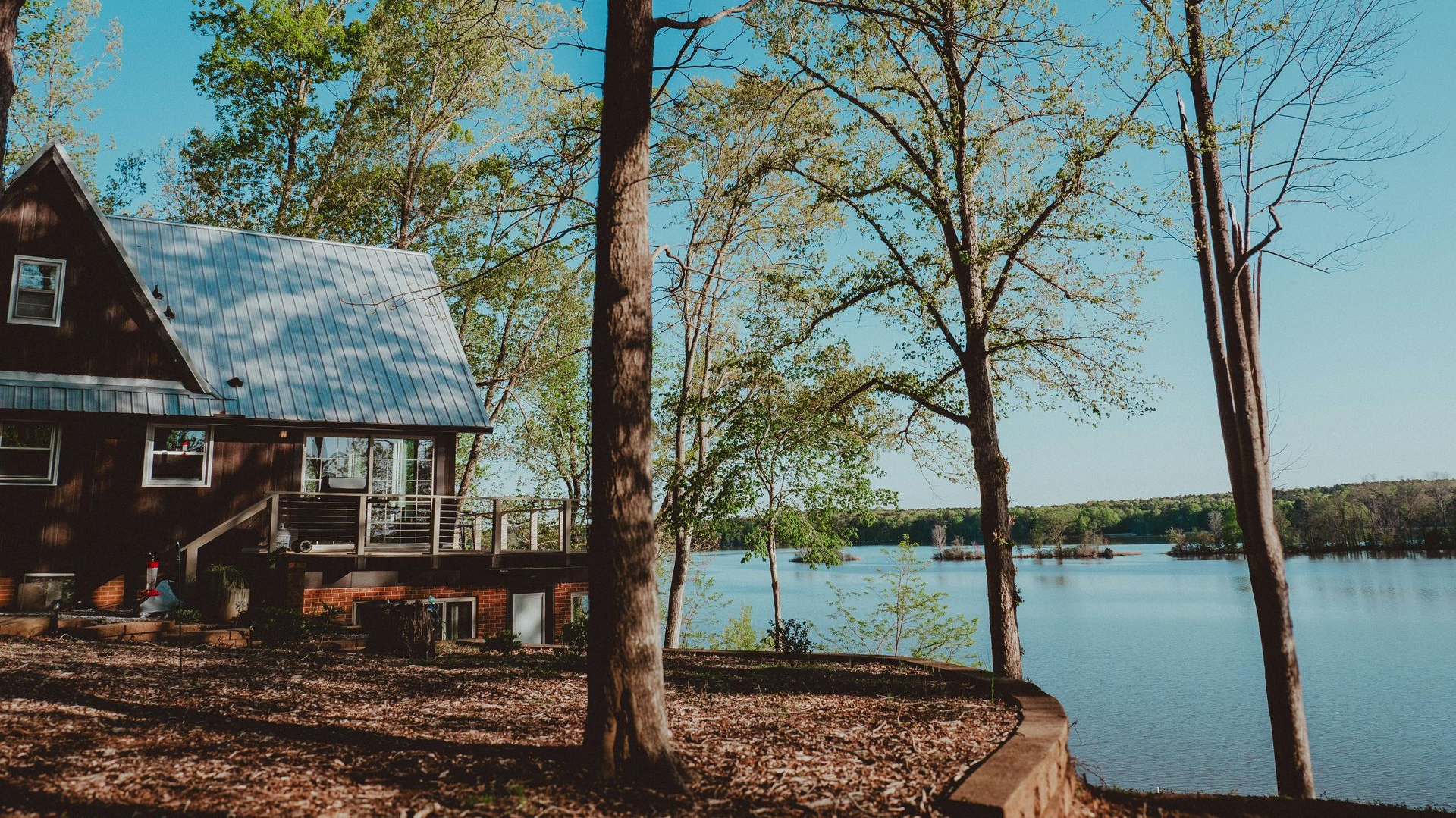 Lakeside cabin with tall trees, blue sky, and a calm lake in the background
