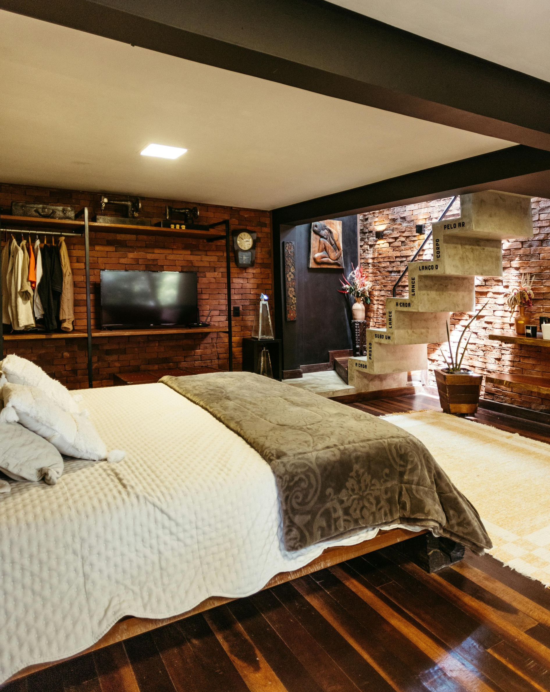 Modern loft bedroom with exposed brick, wooden floors, bed, TV, and open staircase