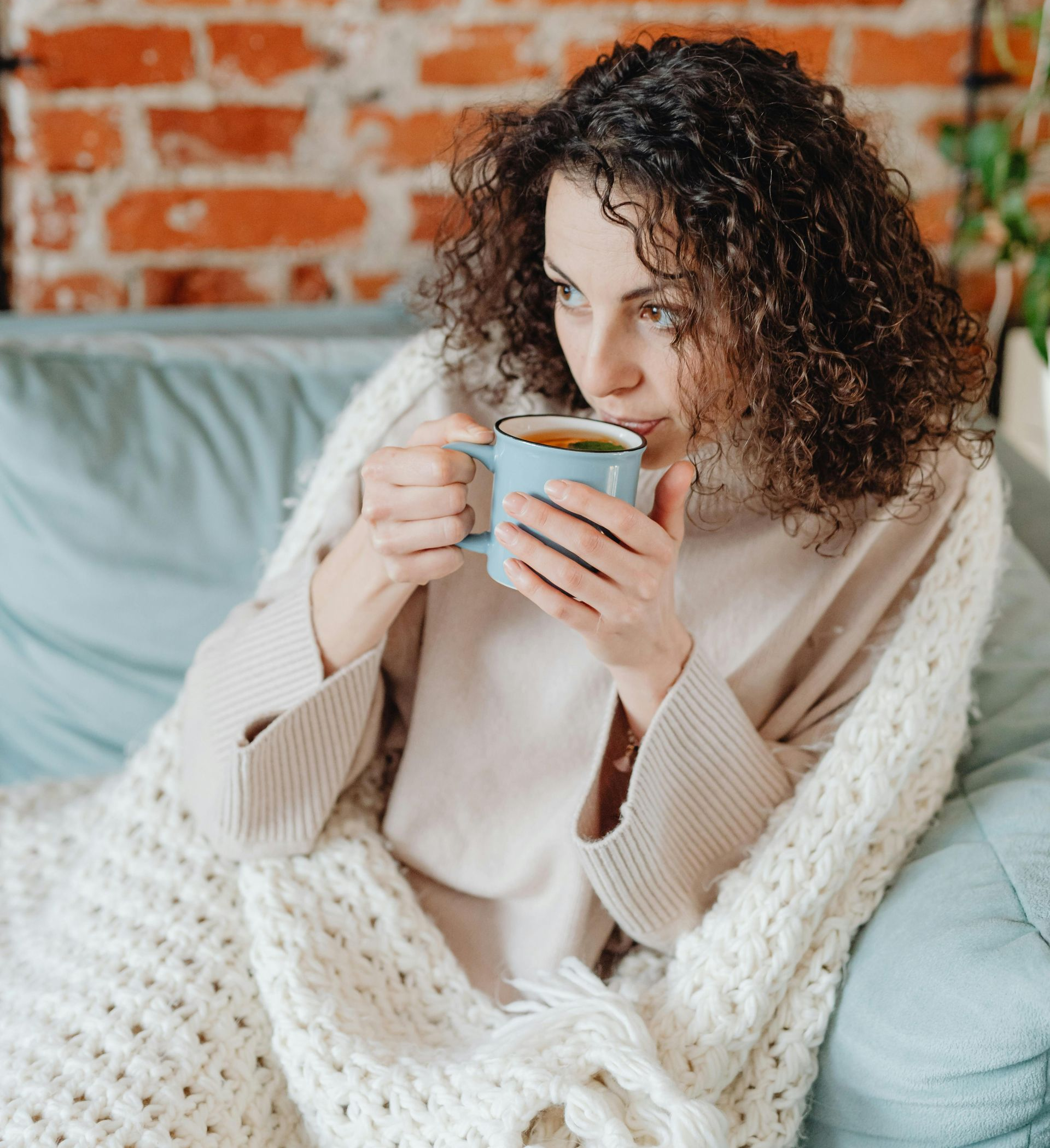 Woman in a cream sweater holding a mug on a sofa, with a brick wall and cozy blanket behind her