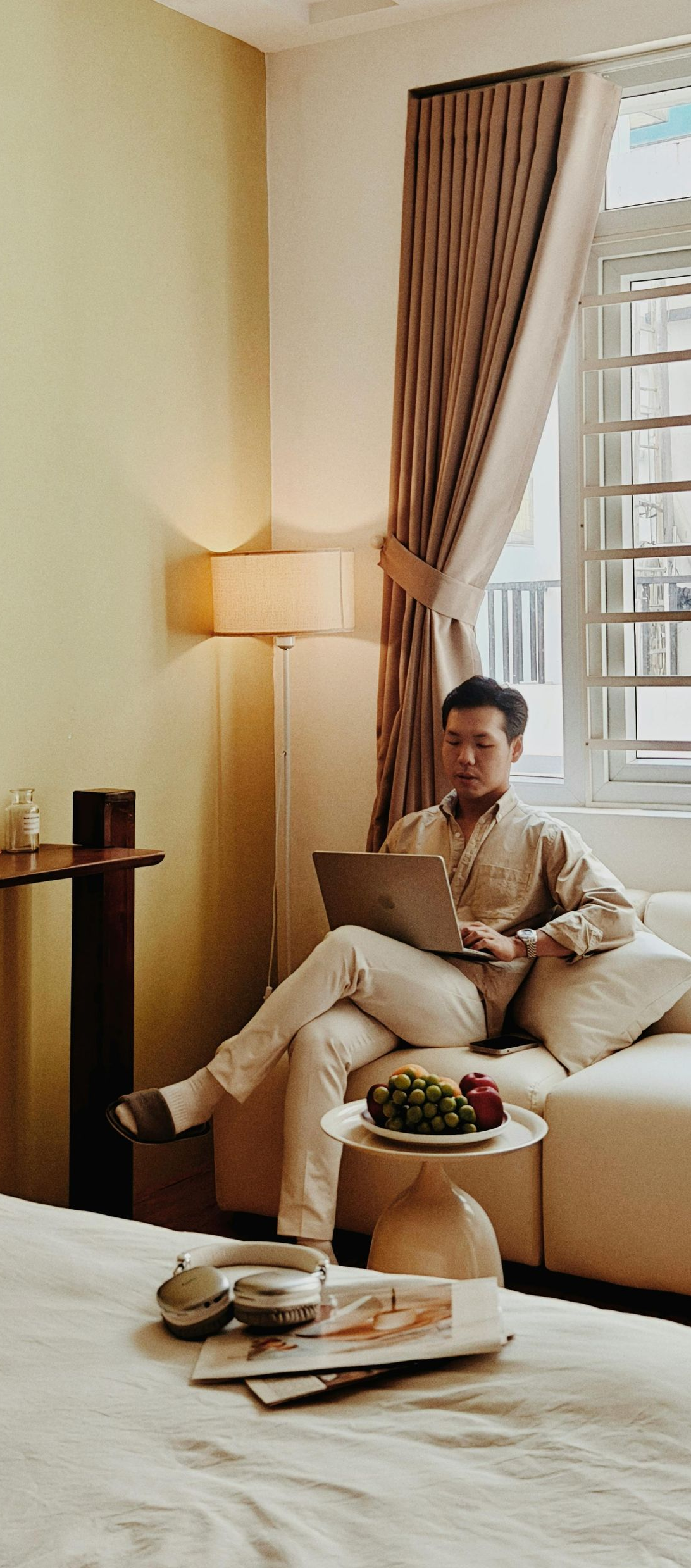 Person relaxing on a sofa in a sunlit living room with curtains, lamp, and a coffee table of snacks