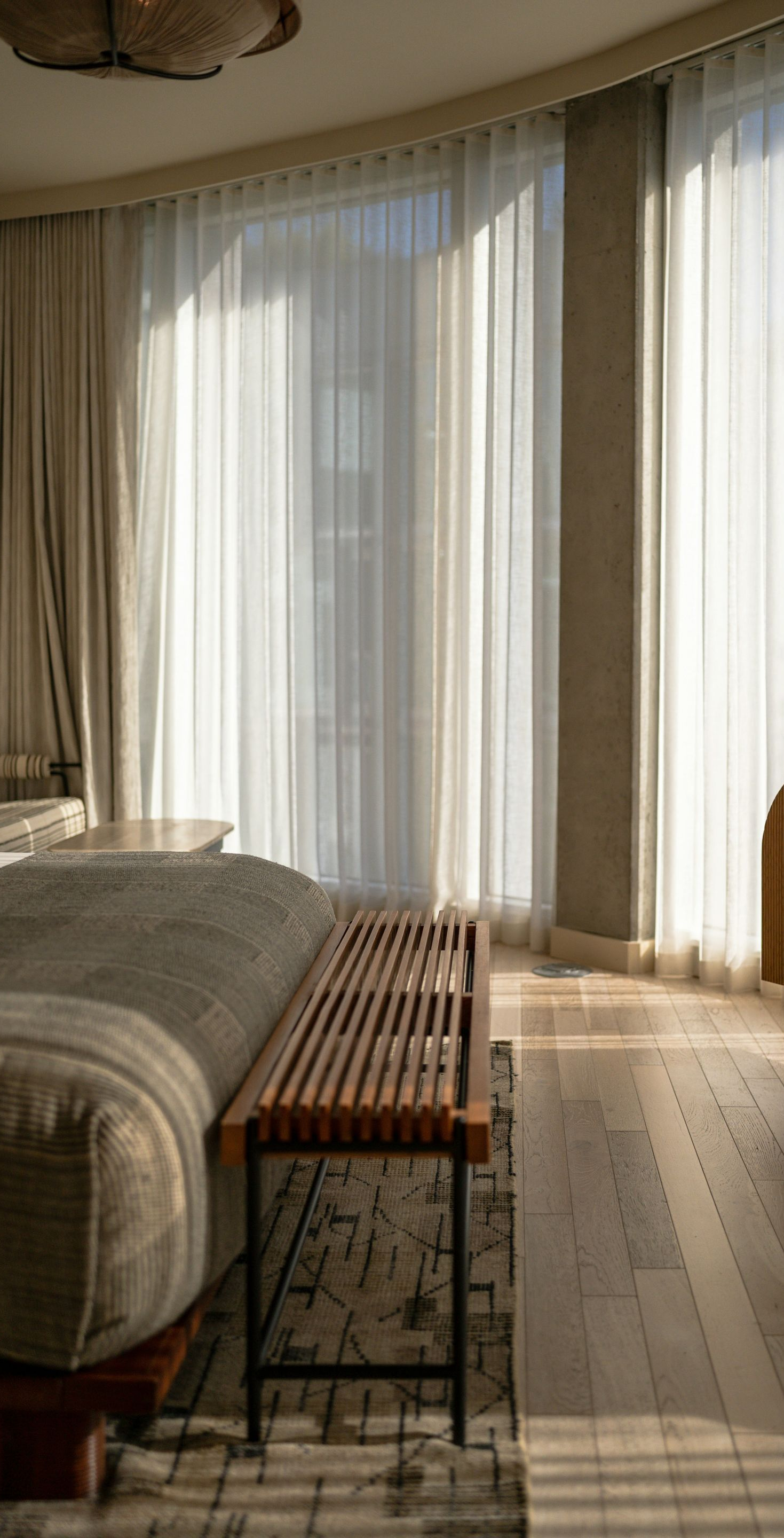 A modern bedroom featuring a wood slat bench at the foot of a bed, large windows with sheer curtains, and wooden flooring.