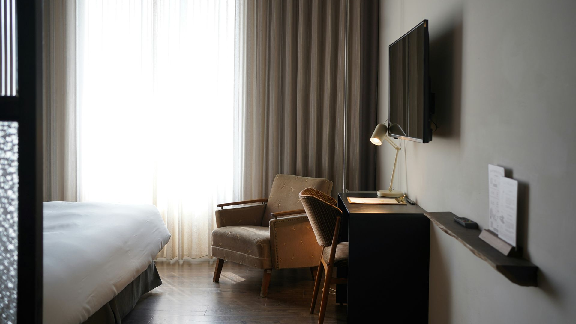 A modern hotel room with a bed, a beige armchair by a large window with sheer curtains, and a desk with a lamp.