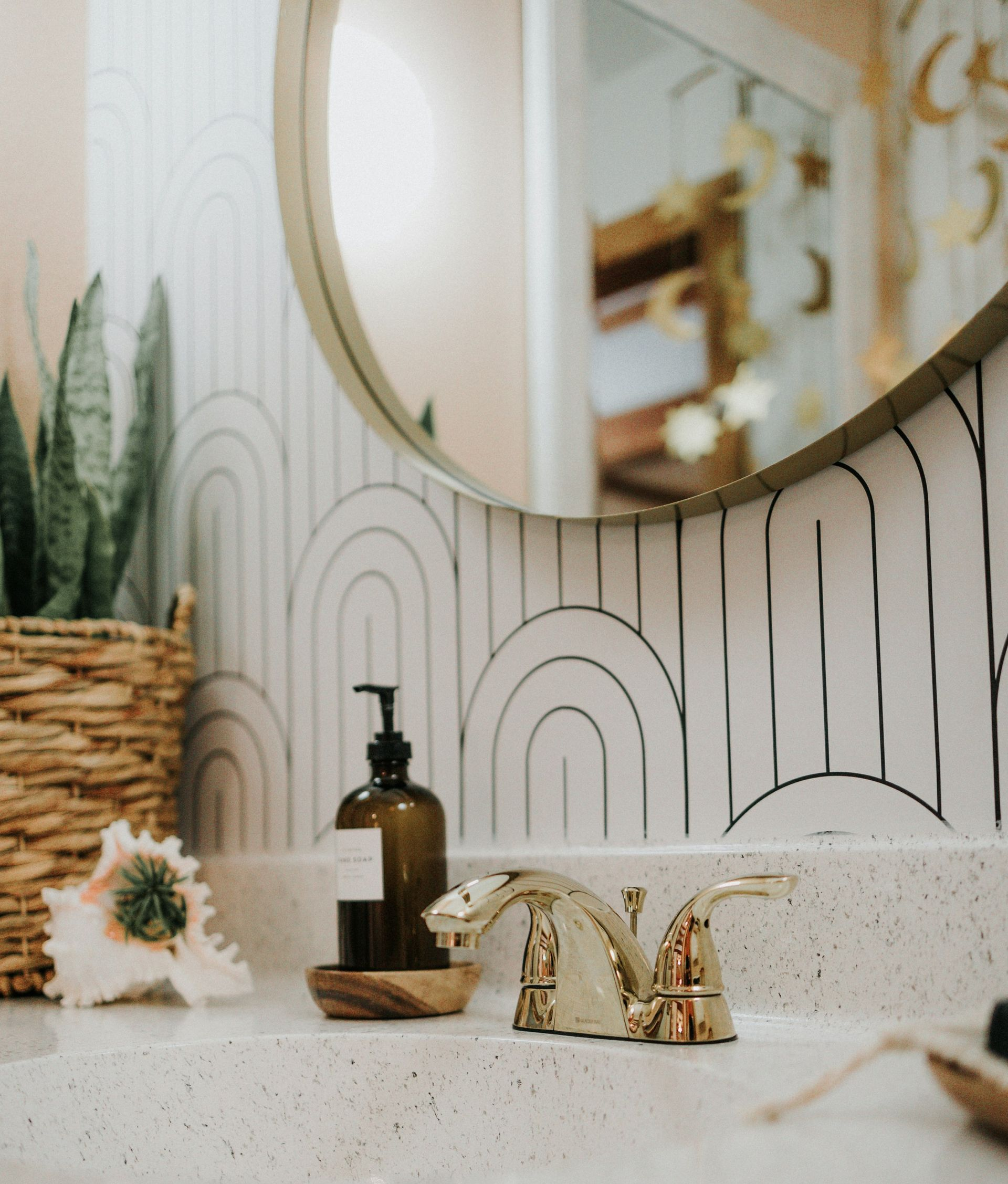 Bathroom sink with gold faucet, round mirror, and patterned tile. Includes a basket with a plant and a seashell.