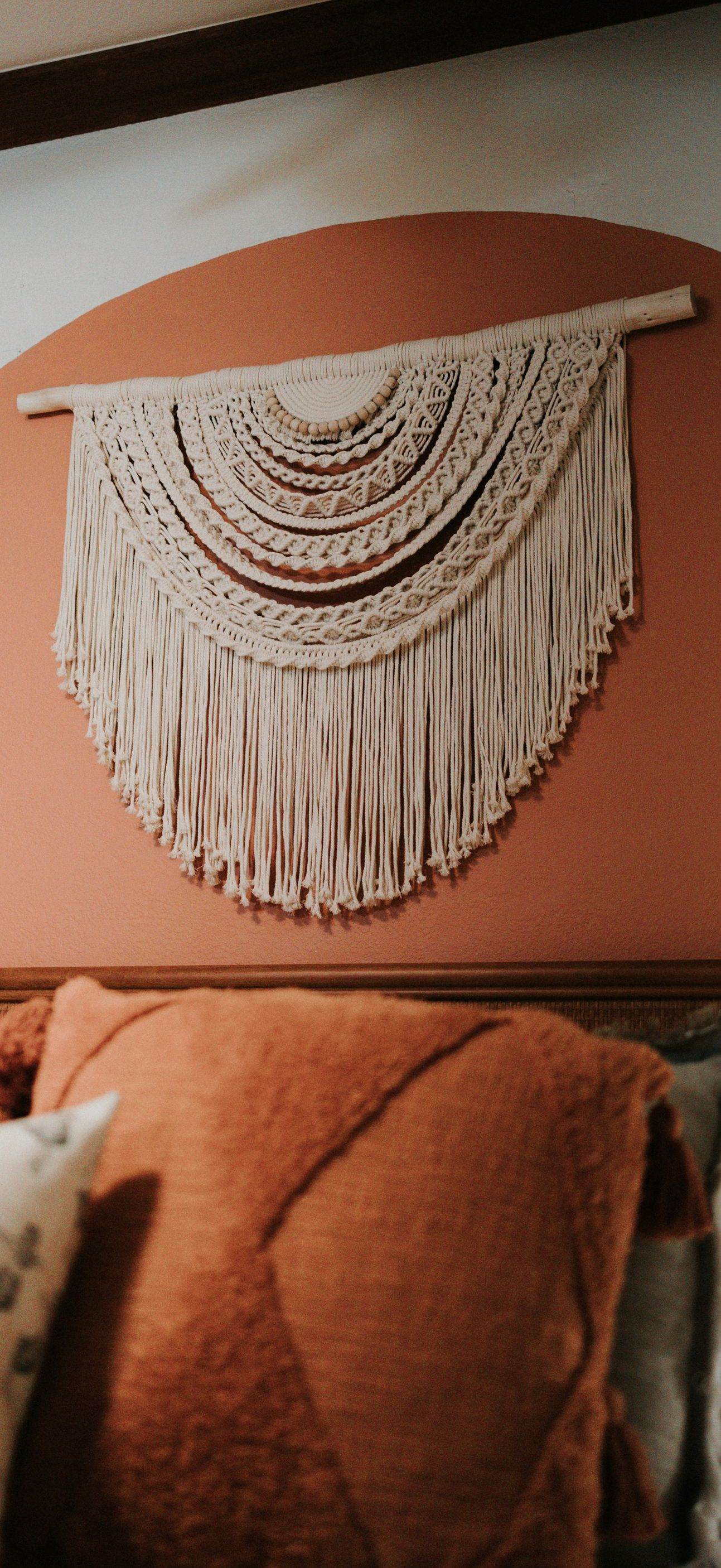 A cream-colored macramé wall hanging mounted above a bed with terracotta-colored pillows against an arched painted wall.
