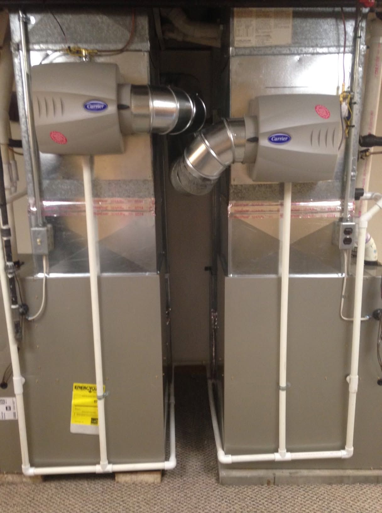 Two gray HVAC furnace units connected by ductwork in a utility room