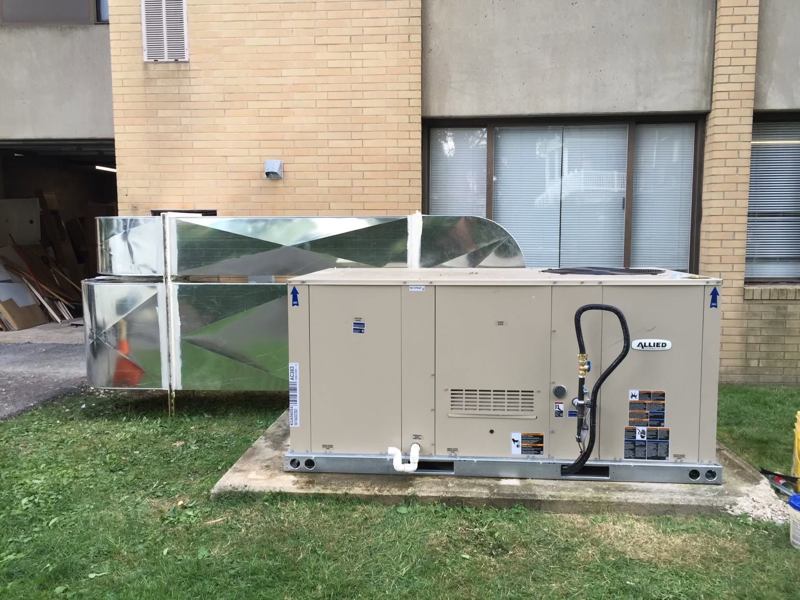 Outdoor HVAC equipment beside a beige building, with metal ducting and a fenced enclosure on grass.