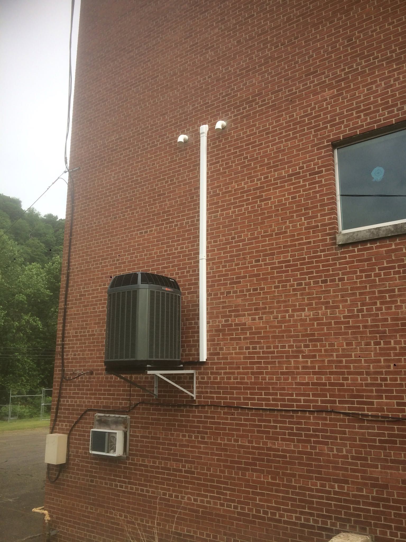 Brick building wall with mounted air conditioner, white pipe, and small windows.