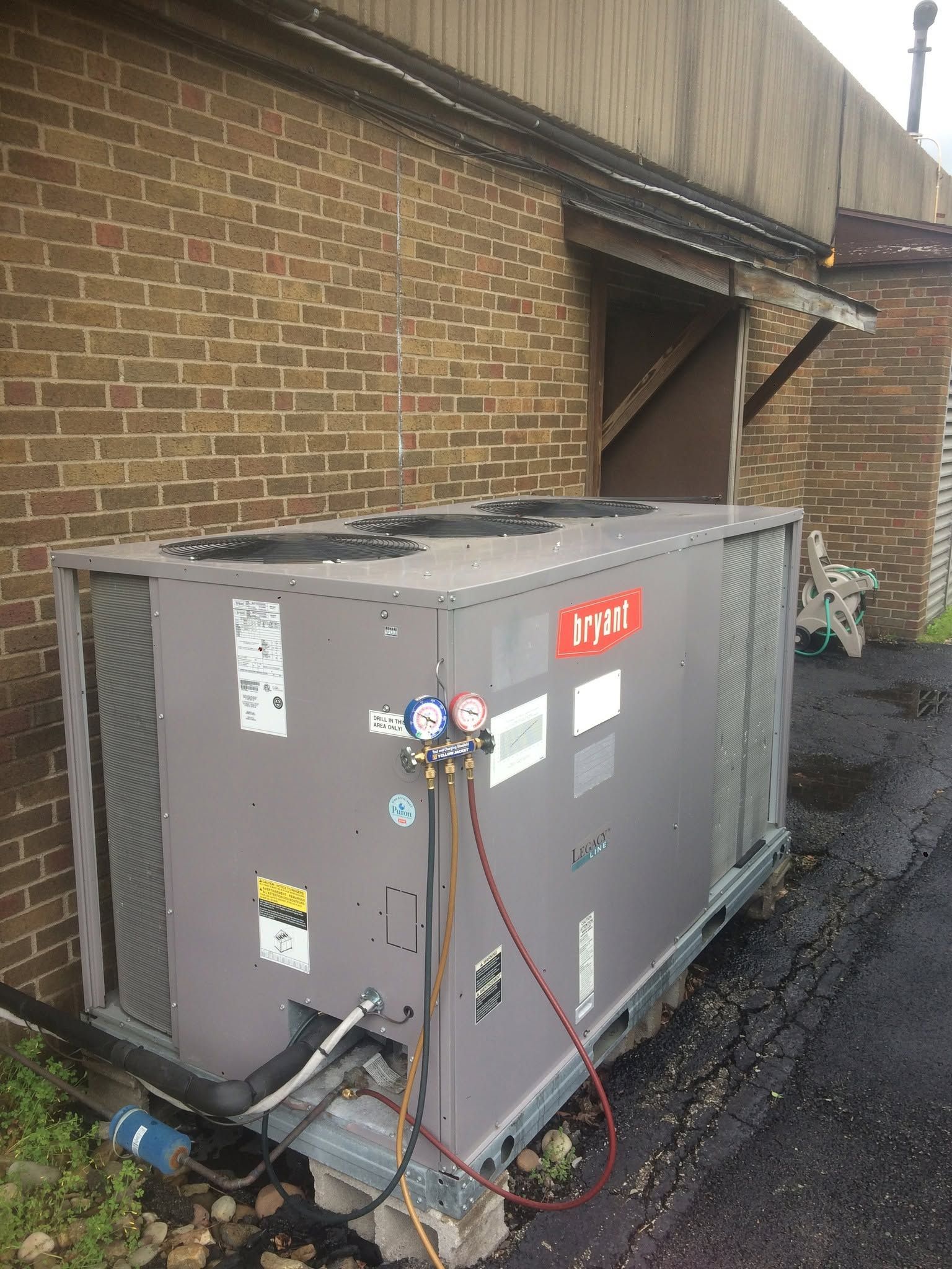 Large gray HVAC unit beside a brick wall, with hoses and cables attached near a building entrance.