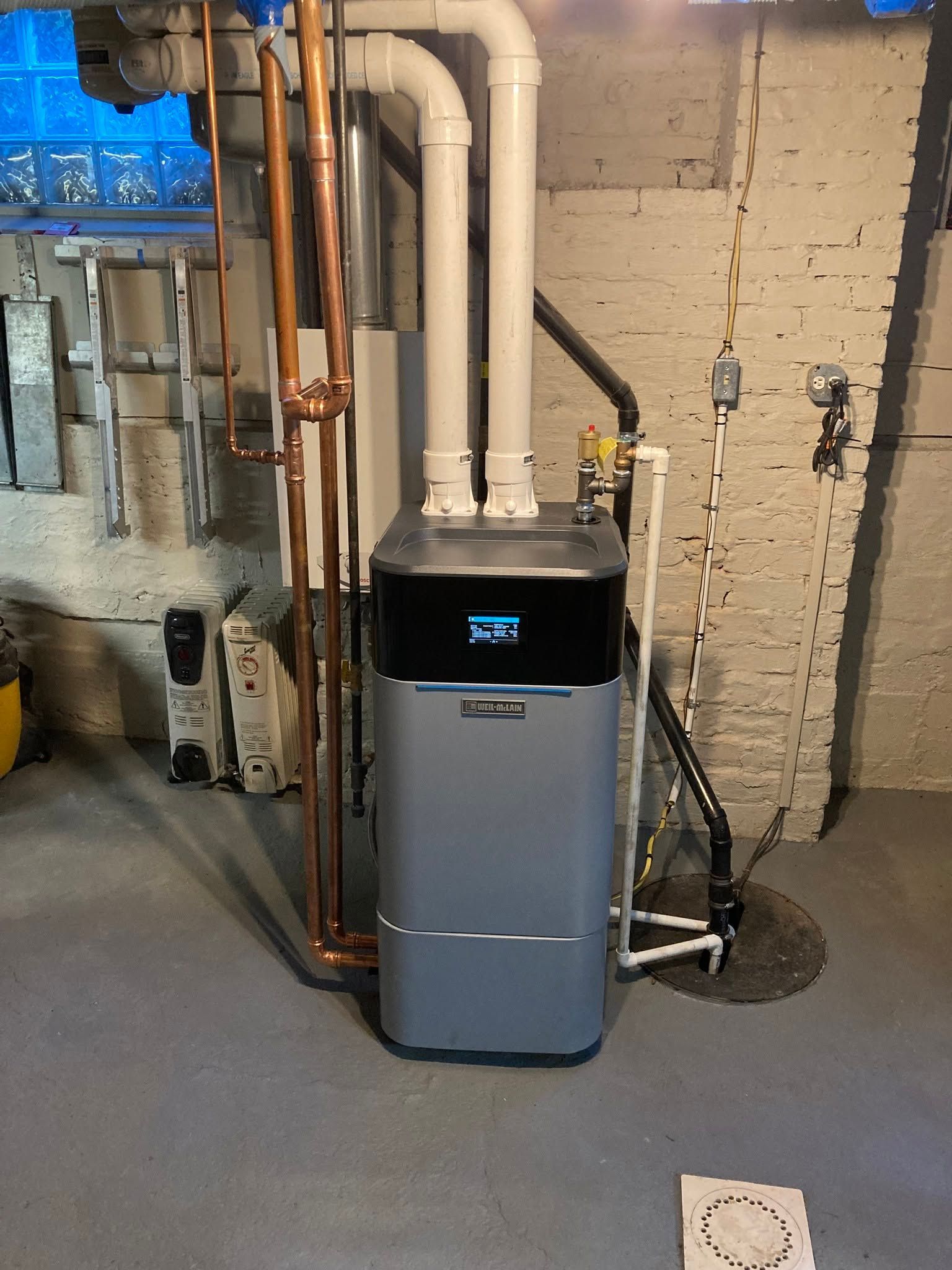 Basement utility room with a gray boiler or furnace beside exposed copper pipes and brick wall.