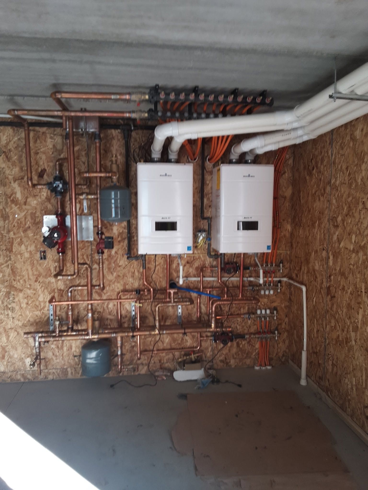 Utility room with two white wall-mounted boilers and exposed copper plumbing on a wood-paneled wall.