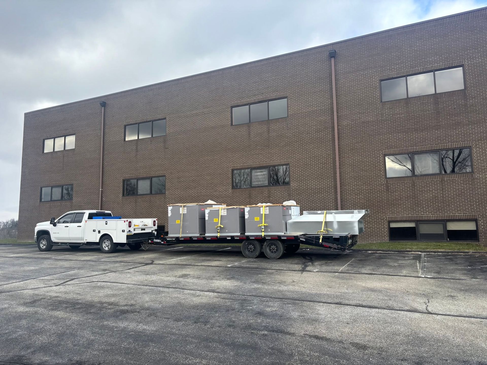 White truck towing a flatbed trailer in front of a brown industrial building on a gravel lot