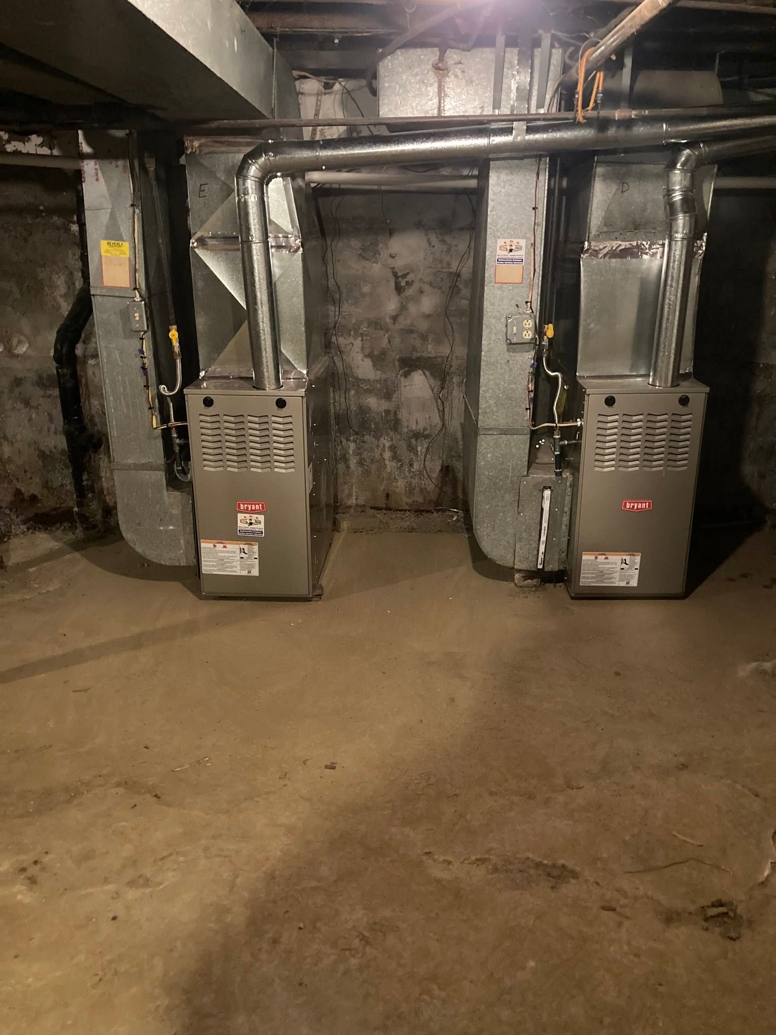 Two gray furnaces in a dim basement with exposed pipes and ducts.