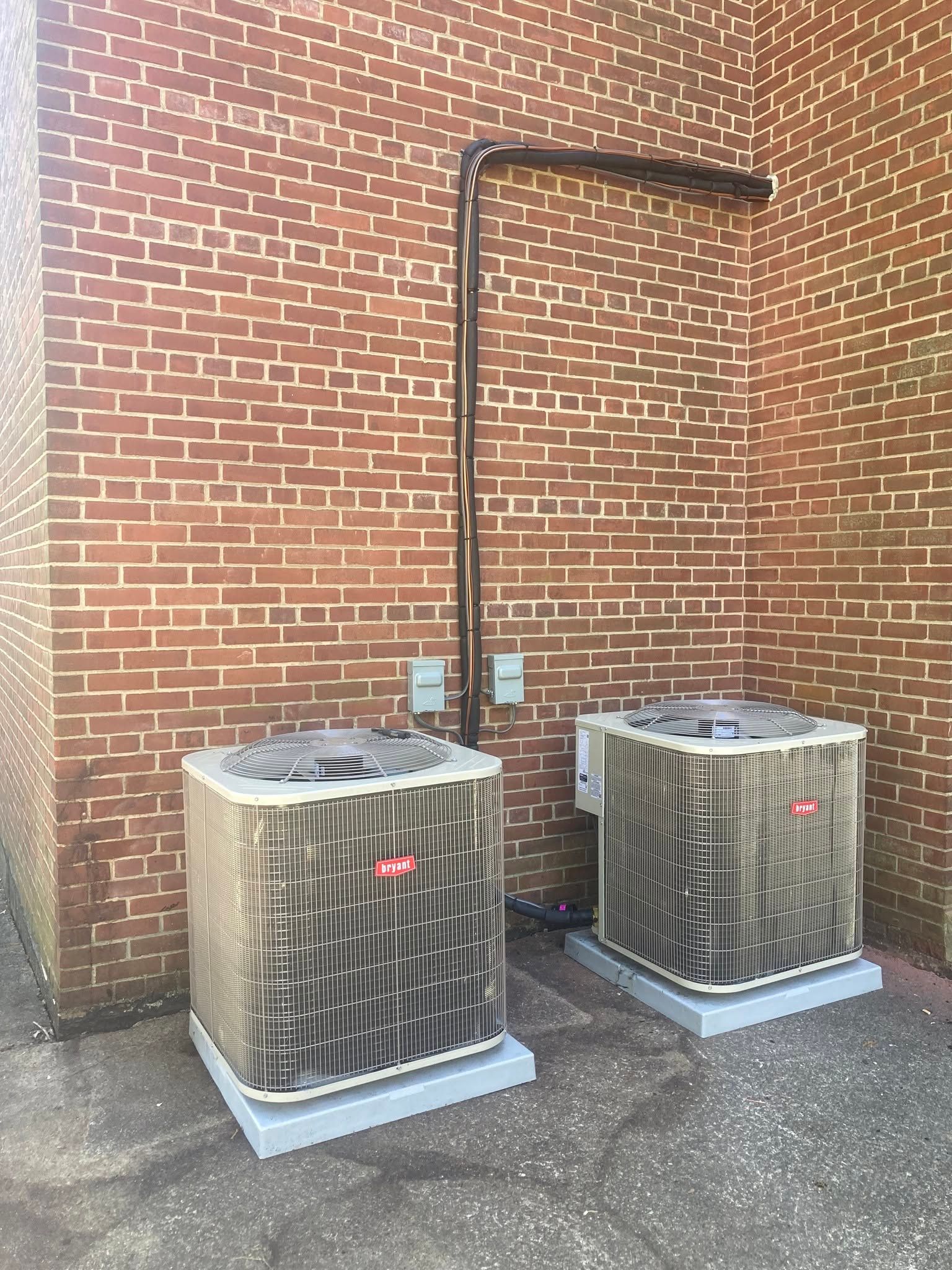 Two outdoor HVAC units against a brick wall with a copper pipe rising above them