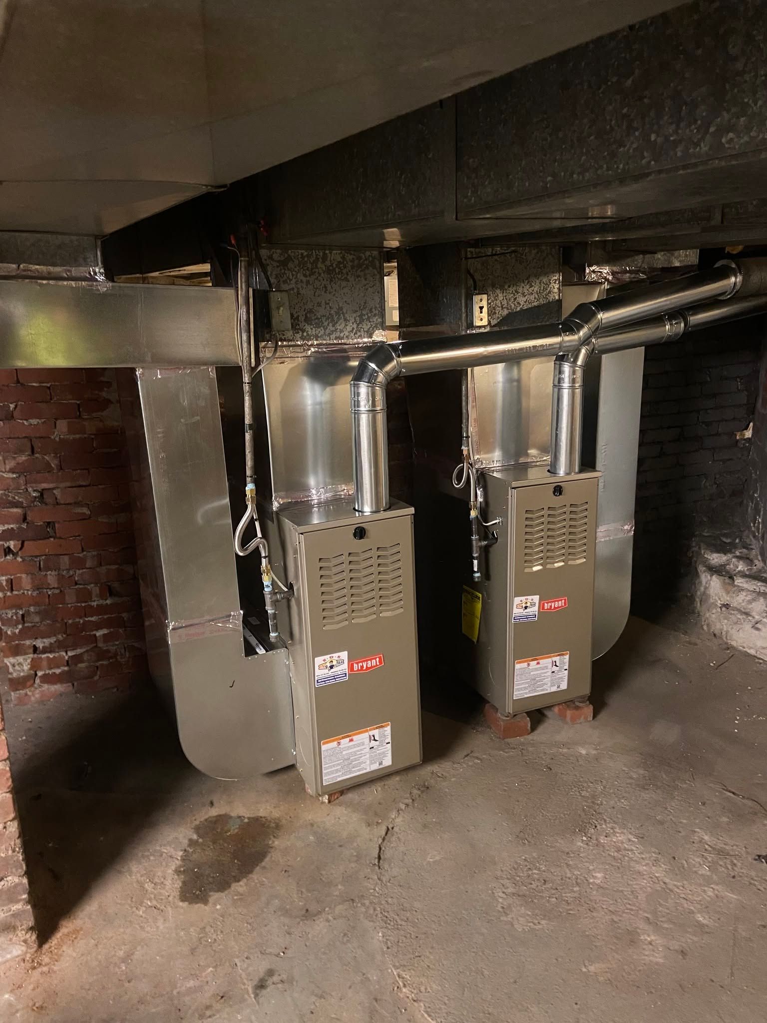 Two gray furnaces with shiny ductwork in a dim basement utility room.