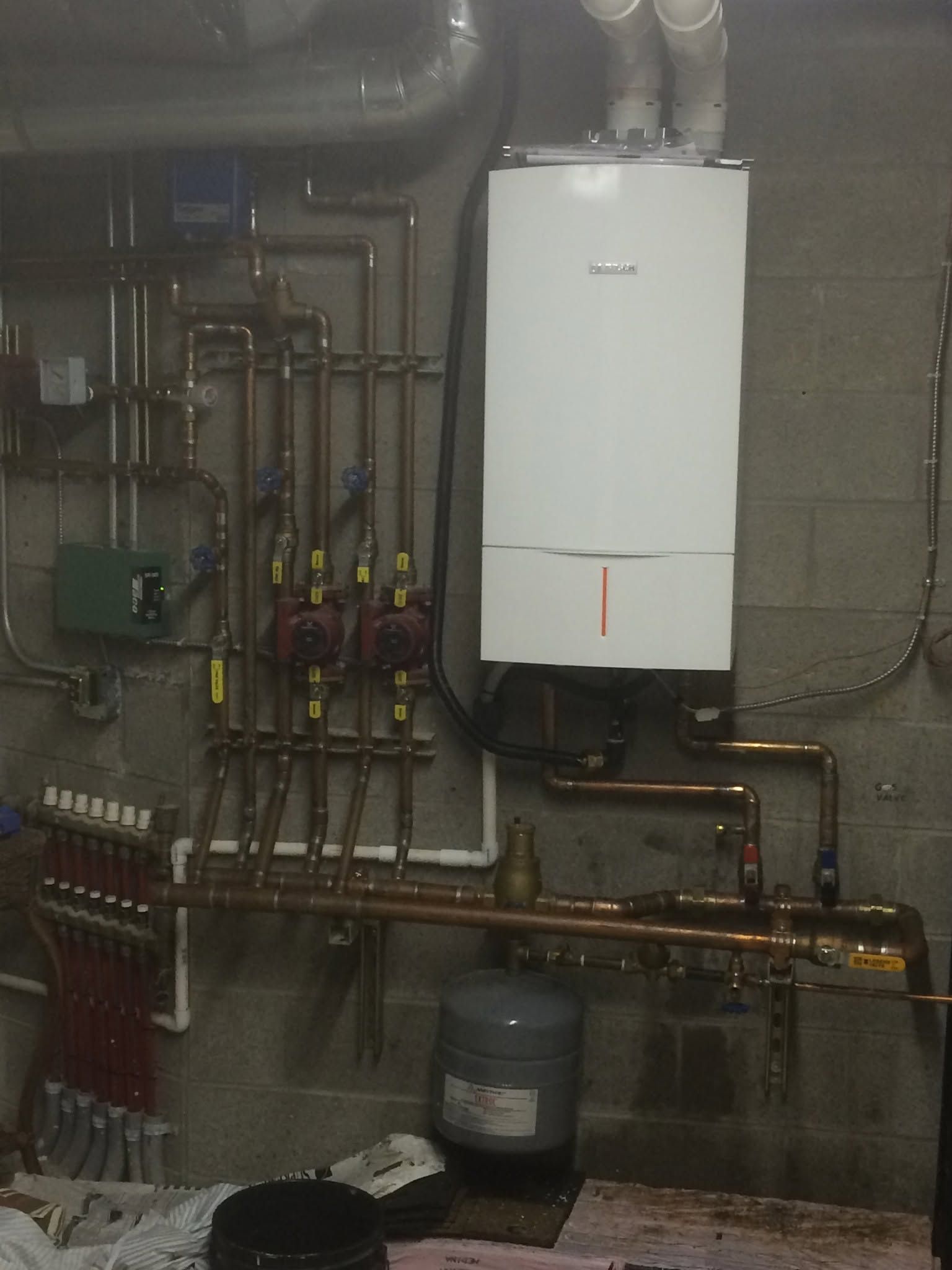 Boiler room wall with white boiler, exposed pipes, valves, and a green control box on a cinderblock wall