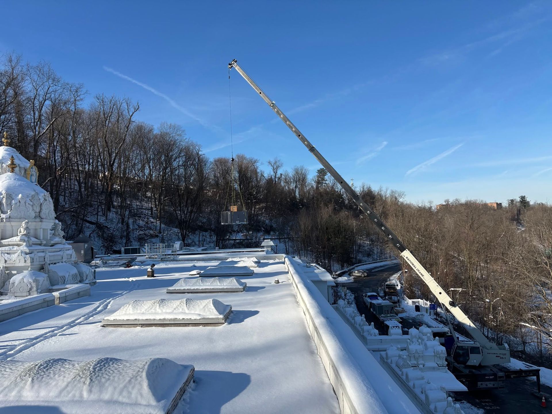 Snow-covered rooftop with a crane lifting materials beside a wooded hillside under a clear blue sky