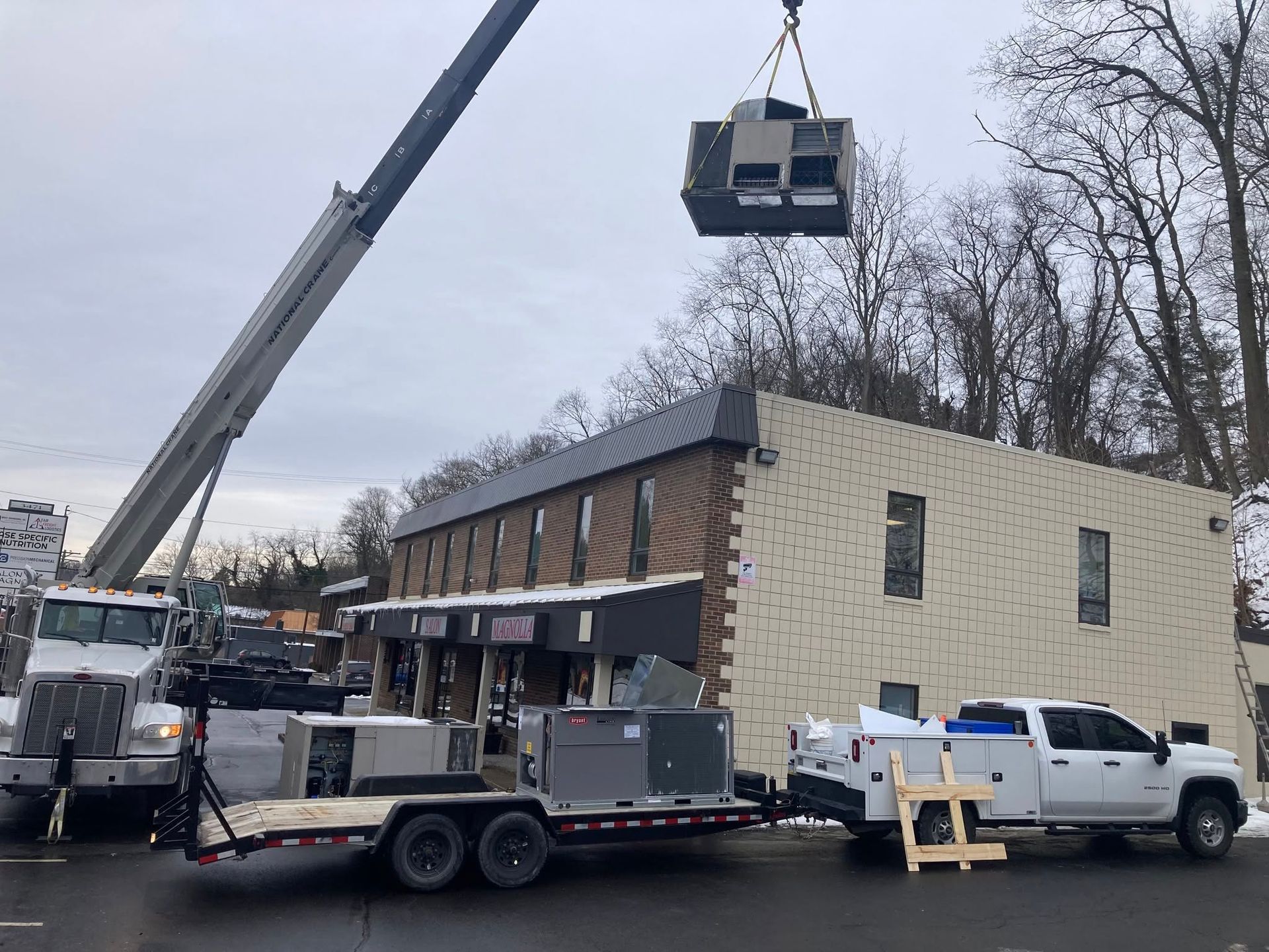 Construction crane lifting a large unit onto a two-story building beside parked work trucks