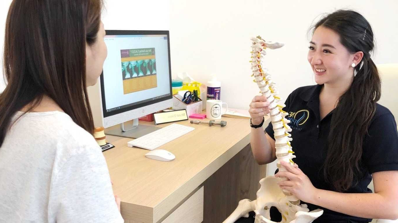 Two people in a clinic; one holds a spine model while discussing X-ray images on a computer monitor.