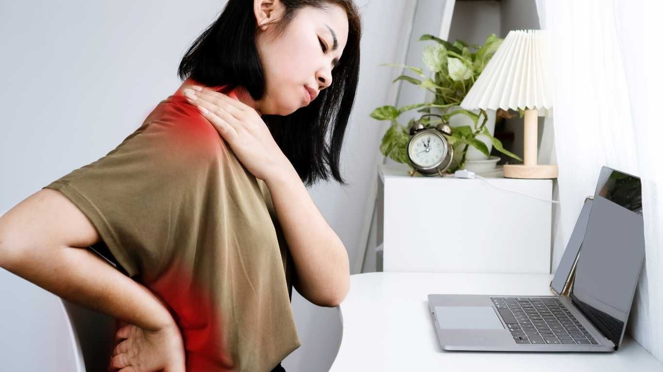 Woman rubbing her shoulder while seated at a desk beside a laptop, looking uncomfortable.
