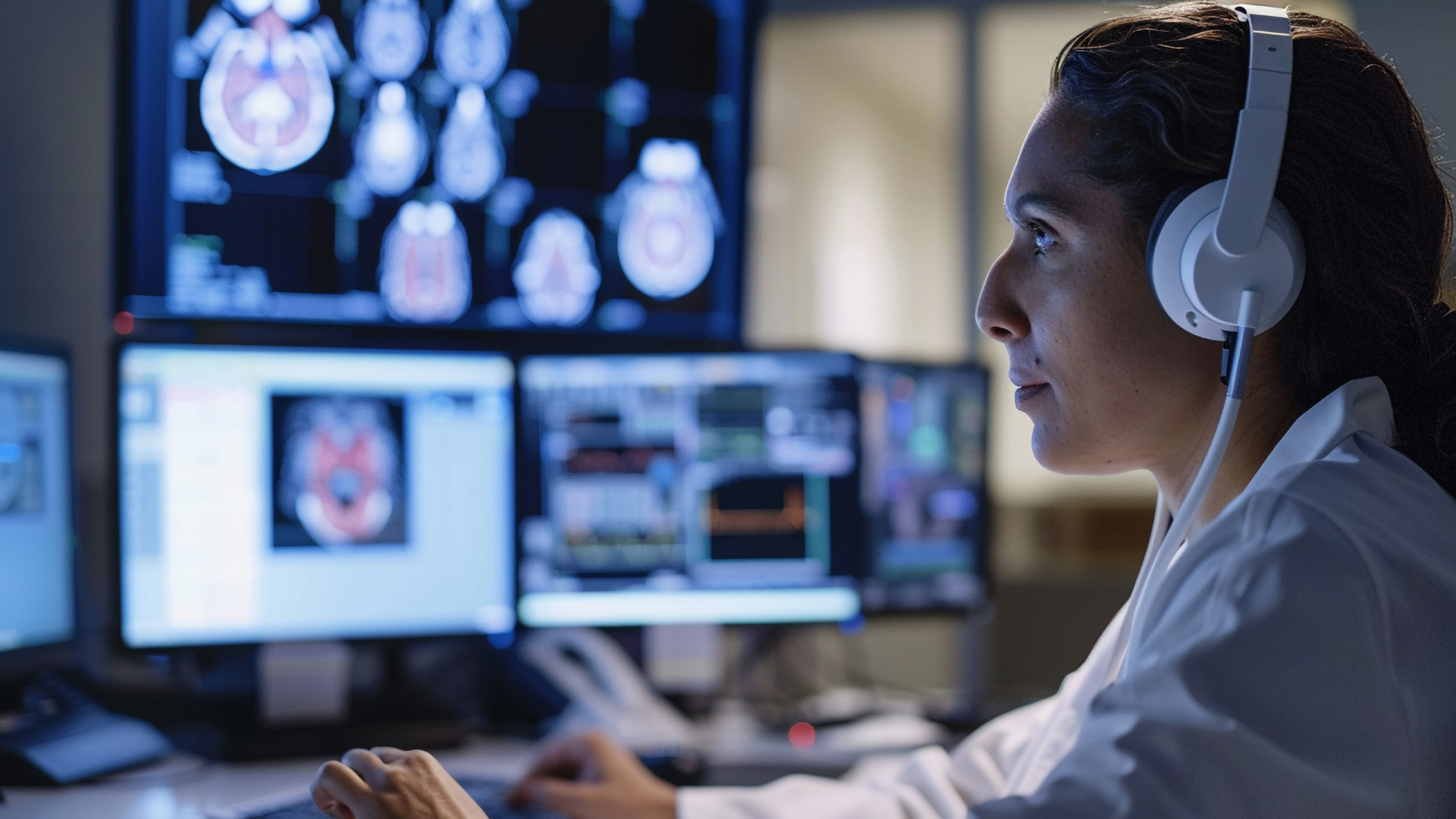 Healthcare worker reviewing brain scans on multiple computer monitors in a dimly lit control room