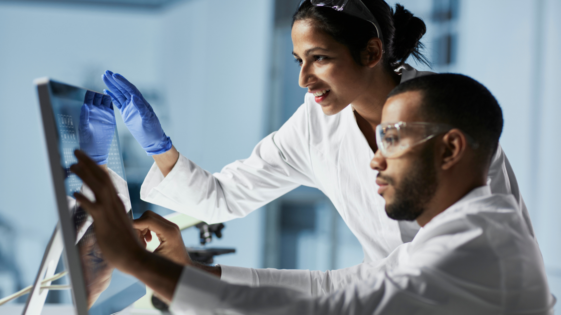 Two lab workers in white coats analyzing data on a computer monitor, one pointing at the screen.