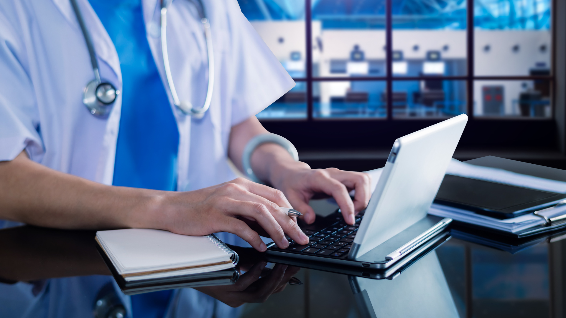 Medical professional typing on a laptop in a clinic, with a stethoscope and notebook on the desk.