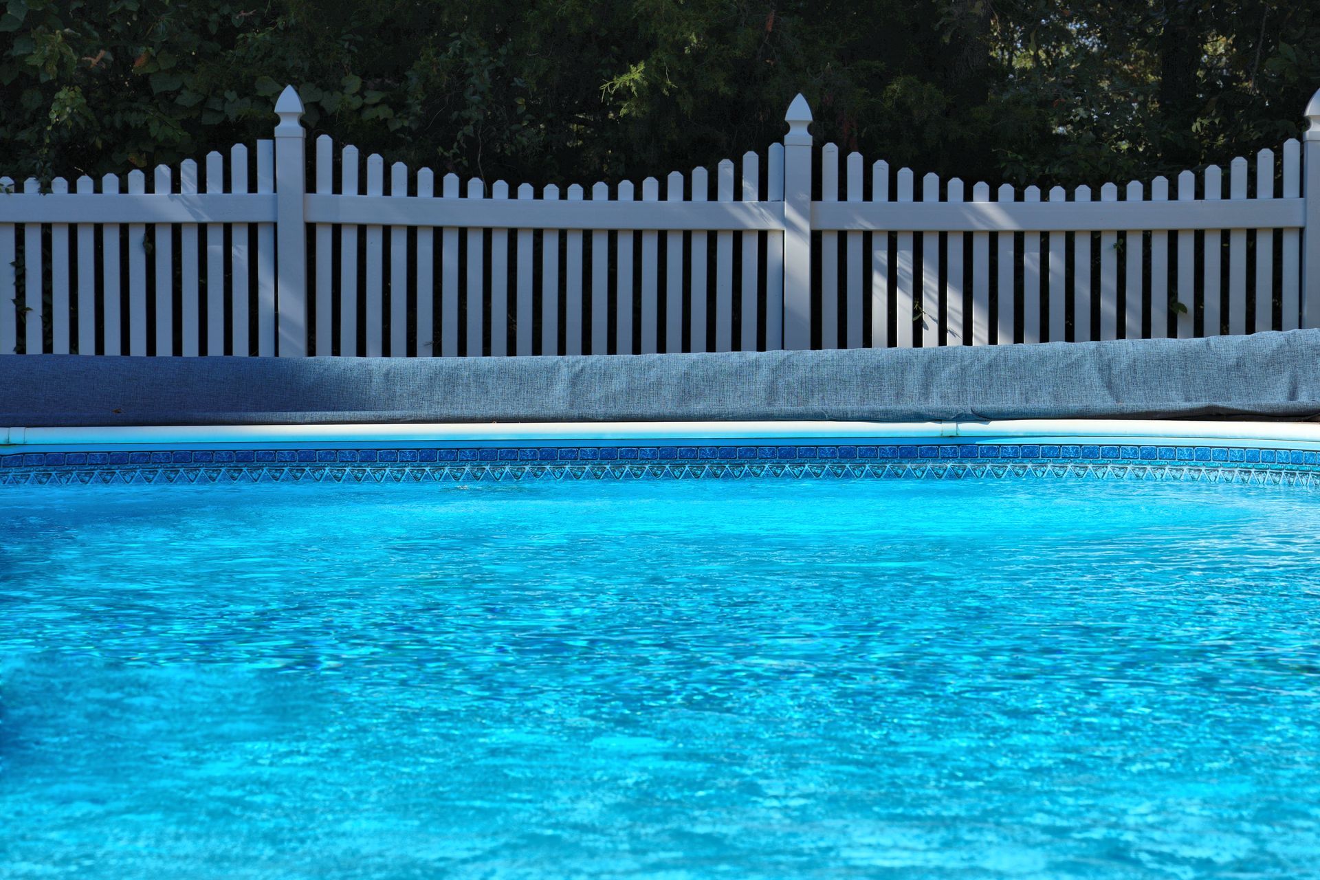 Blue swimming pool water with a white fence and trees in the background