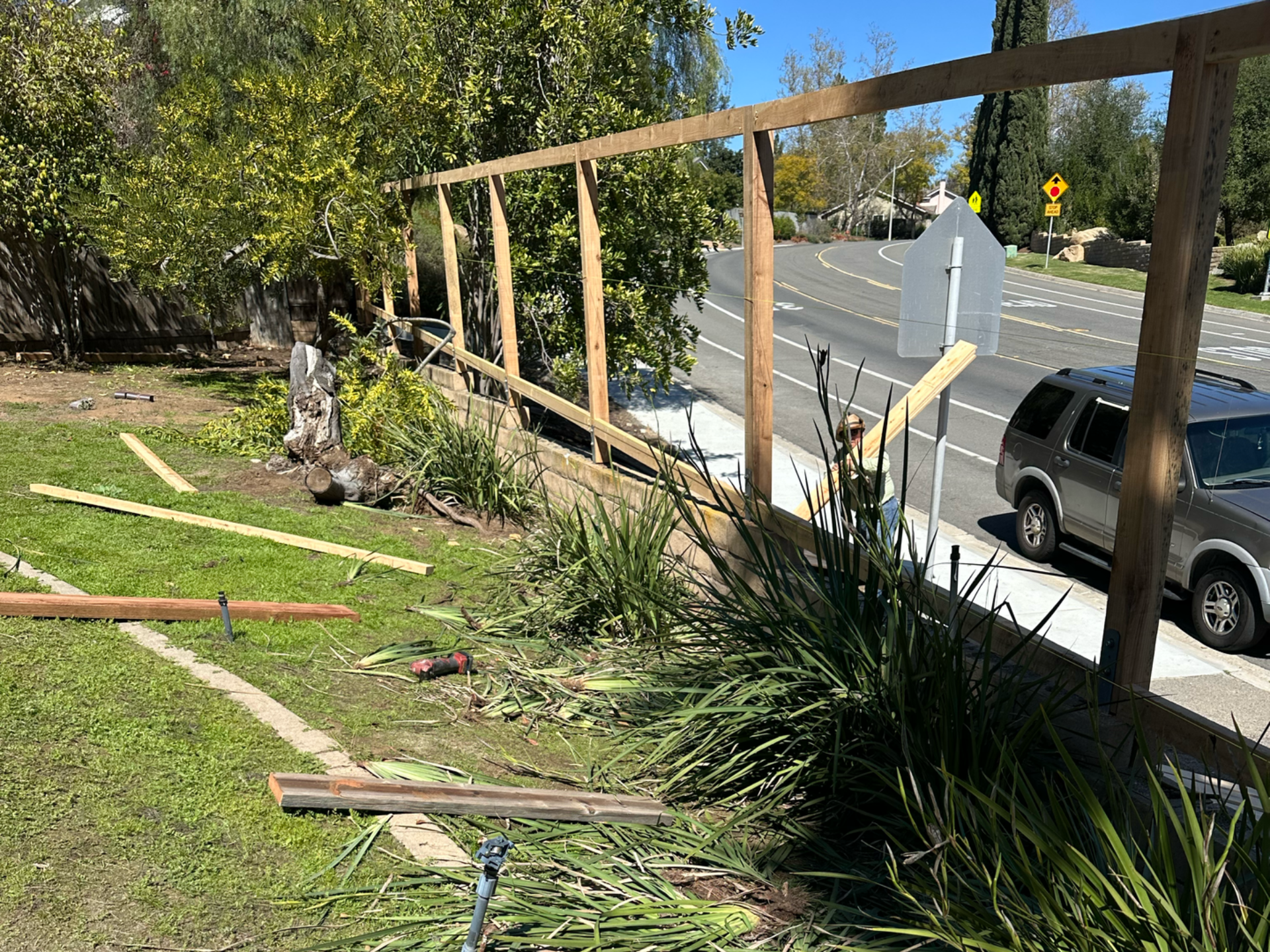 Construction of a wooden fence along a low wall. Green yard, road, and car visible.
