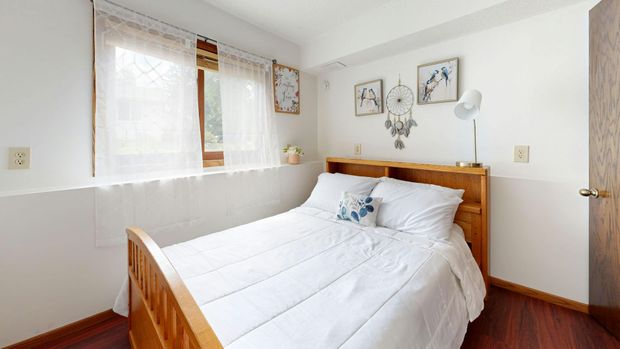 Bedroom with a white bed, wooden headboard, small window with curtains, and wall art.