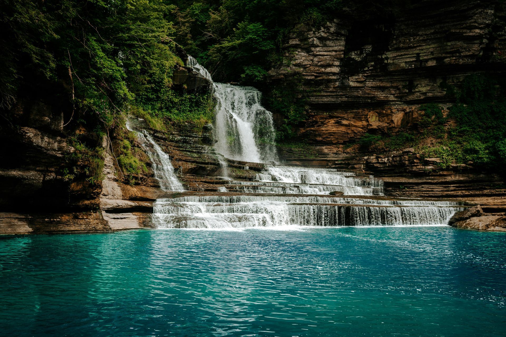 Tiered waterfall spilling into a turquoise pool beneath a rocky cliff and green foliage