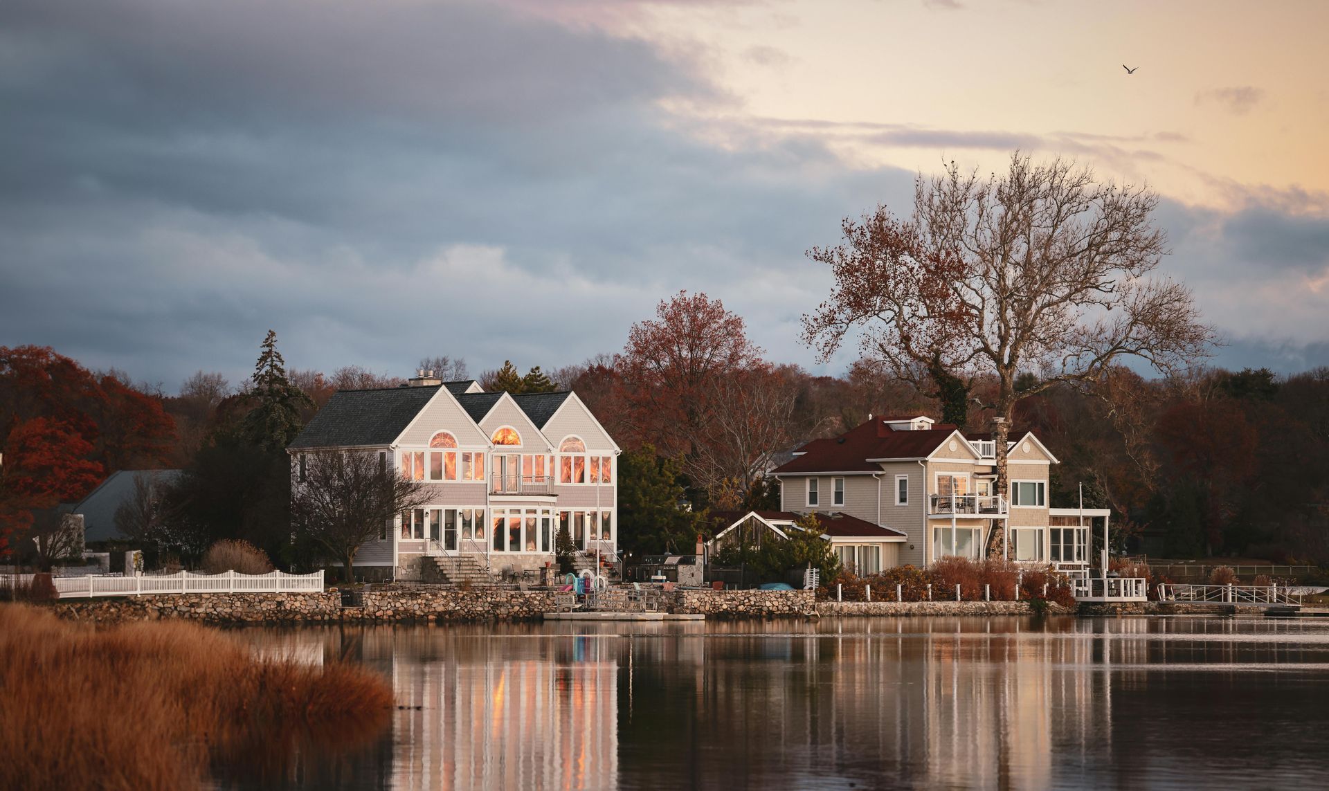 Lakeside houses reflected in calm water at dusk, with autumn trees and a cloudy sky.