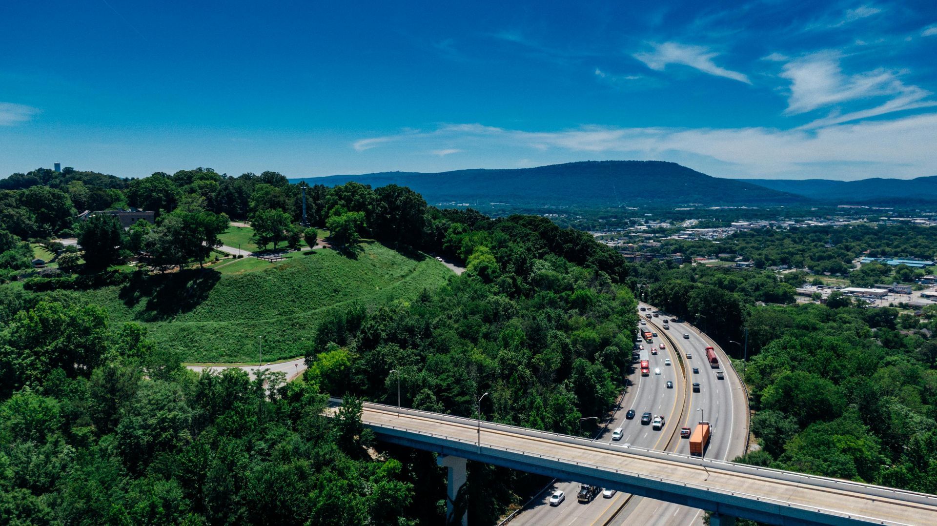 Aerial view of a curving highway beside green hills and trees, with cars driving toward a distant city.