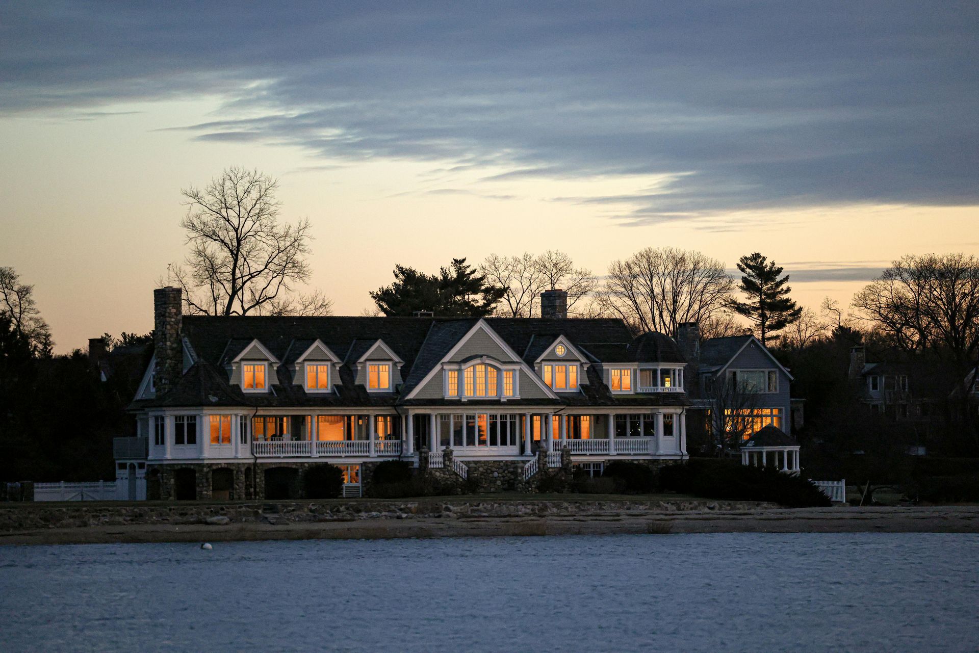 Large white waterfront house at dusk with lit windows and trees silhouetted behind it