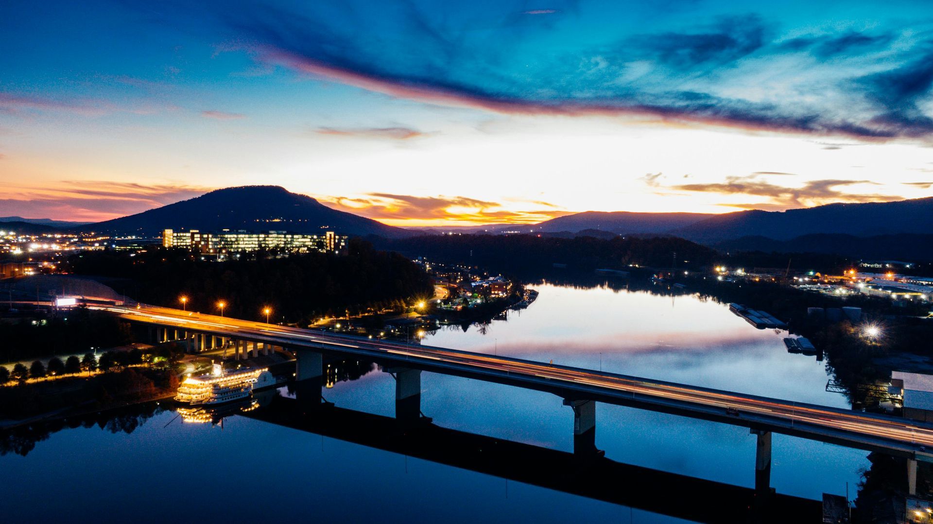 Sunset over a river with a bridge, city lights, and hills in the background