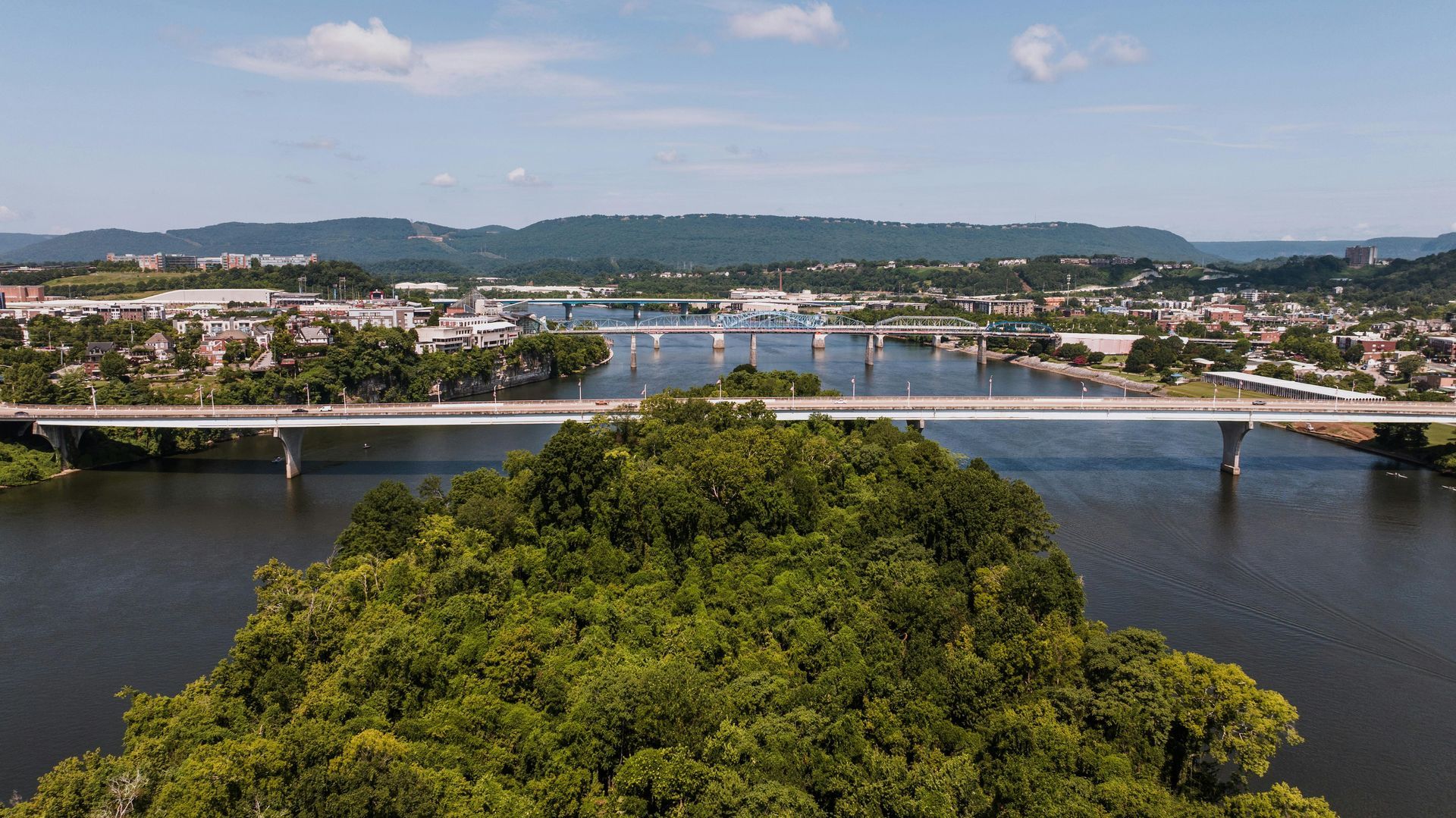 Wide river with a bridge, city buildings, and green trees under a blue sky