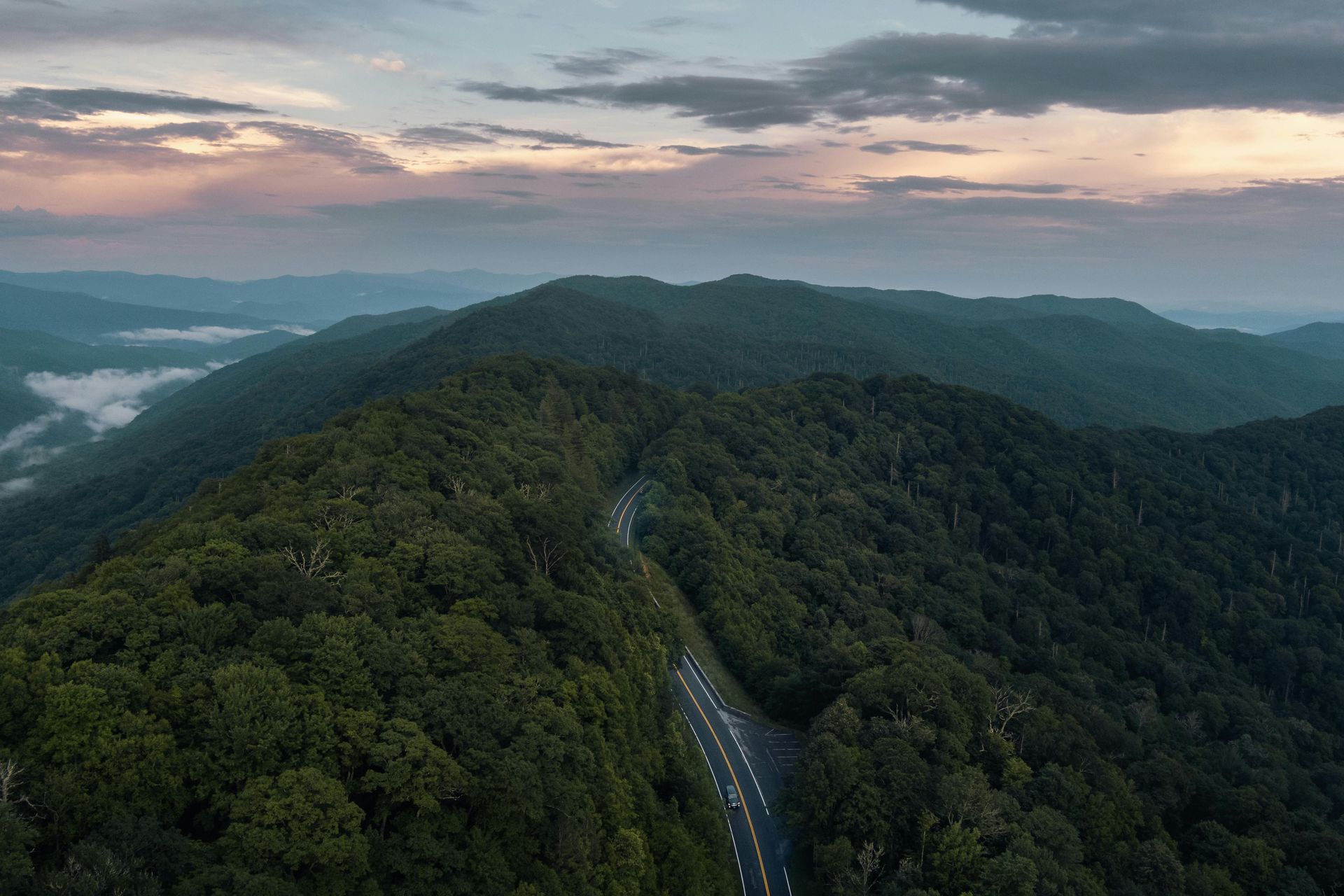 Aerial view of a winding road through dense green mountains at sunrise, with mist over distant hills