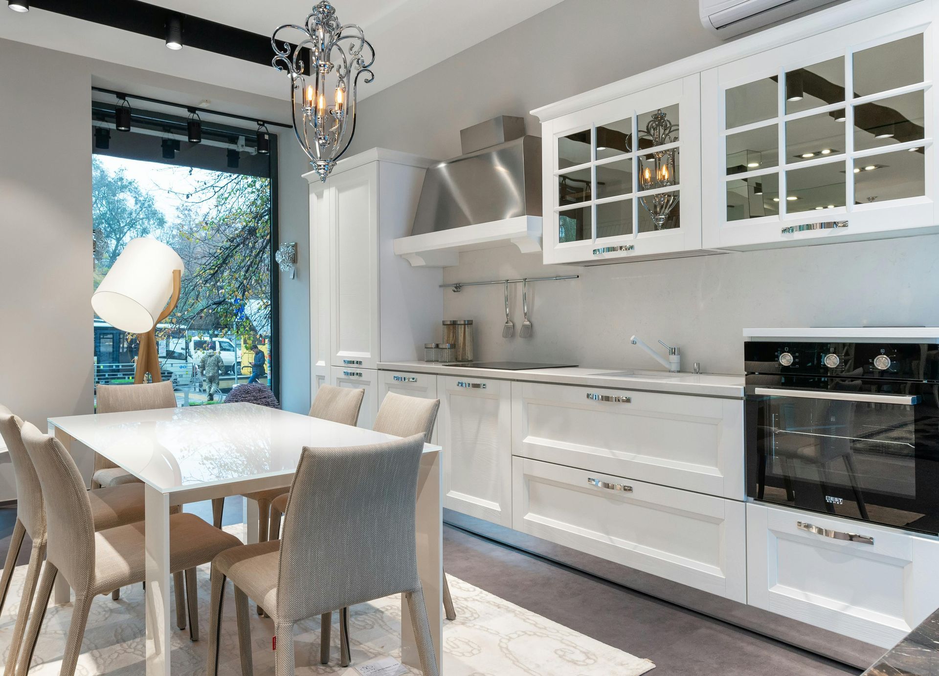 Bright modern white kitchen with dining table, glass-front cabinets, and large window overlooking trees