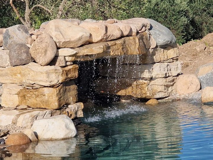 Cascata di pietra che si riversa in una limpida pozza blu tra rocce desertiche e vegetazione.