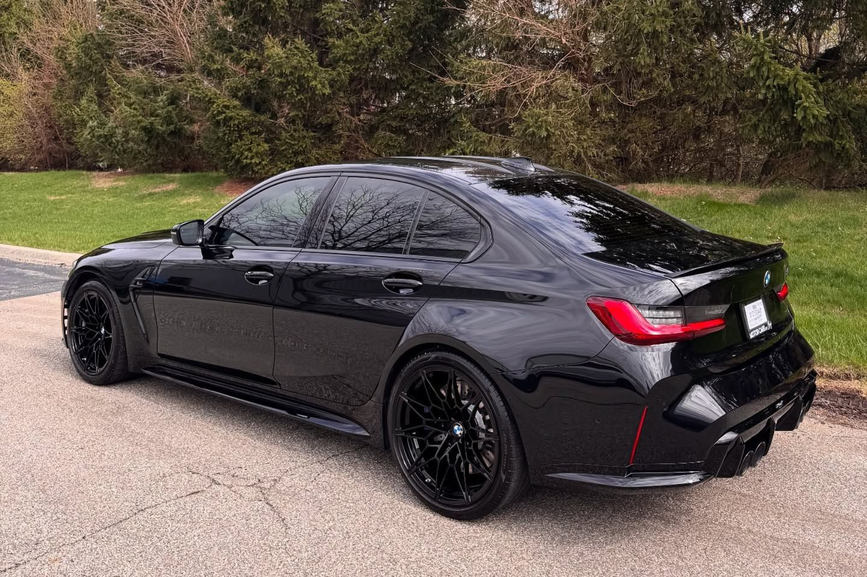 A black BMW M3 sports sedan parked on a paved road next to a grassy area with trees in the background.