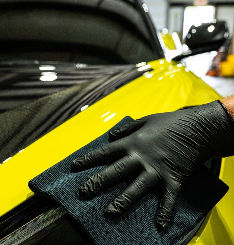 Gloved hand wiping a yellow car with a microfiber cloth; indoor setting.