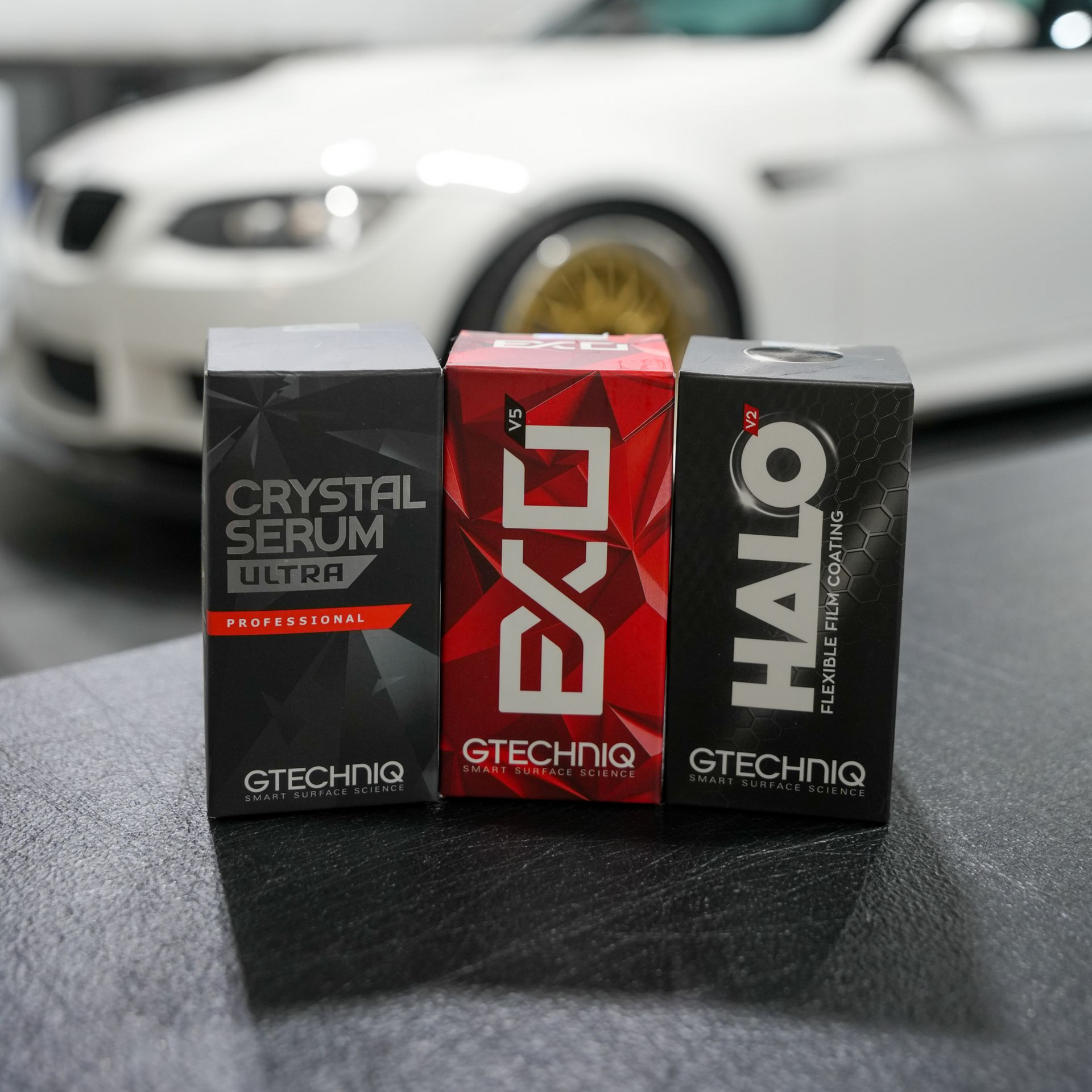 Gtechniq Crystal Serum Ultra, EXO, and Halo ceramic coating bottles lined up in a workshop with a white BMW in the back.