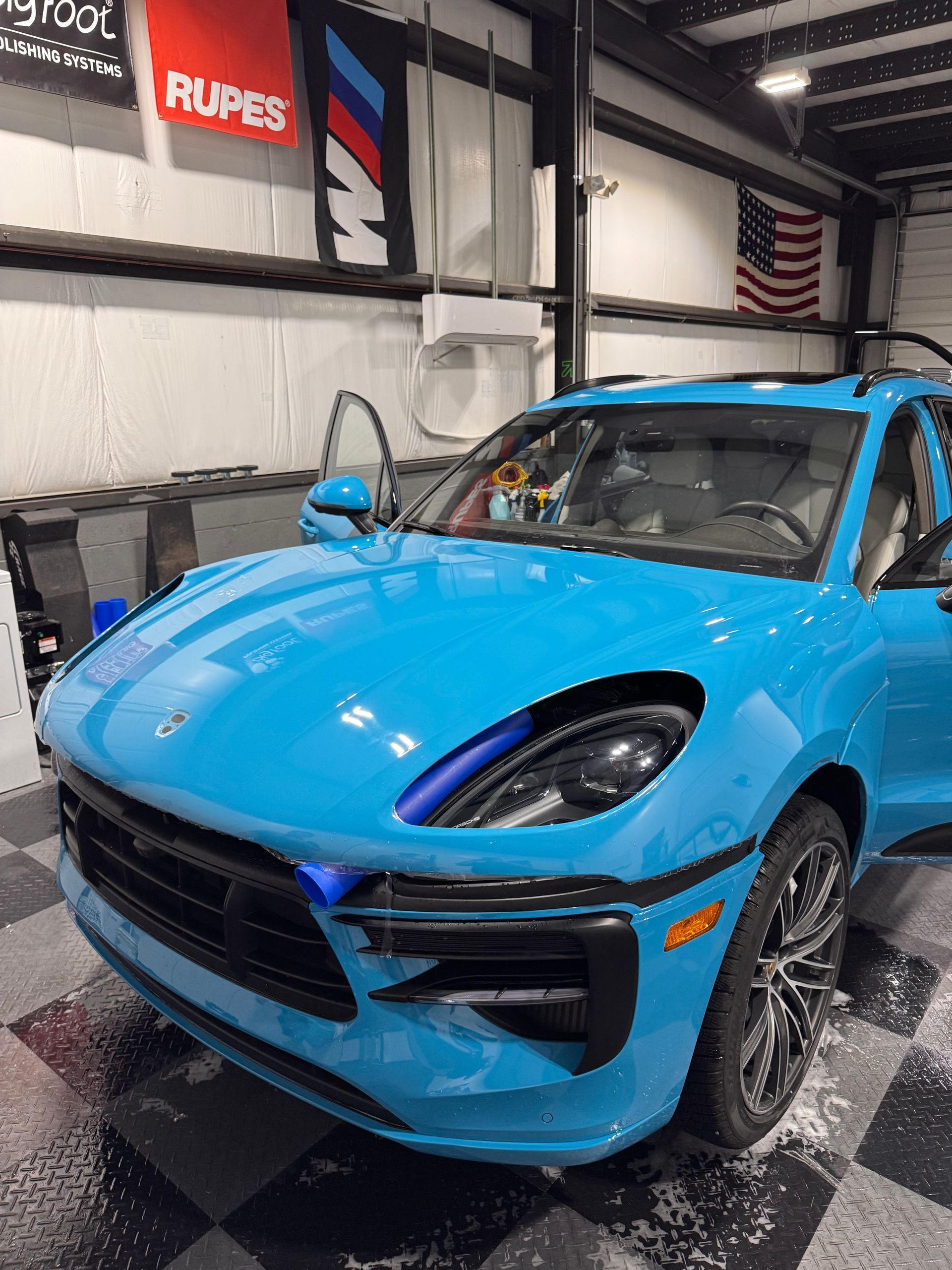 A bright blue Porsche Macan sits in a garage with tiled floors and automotive branding on the walls.
